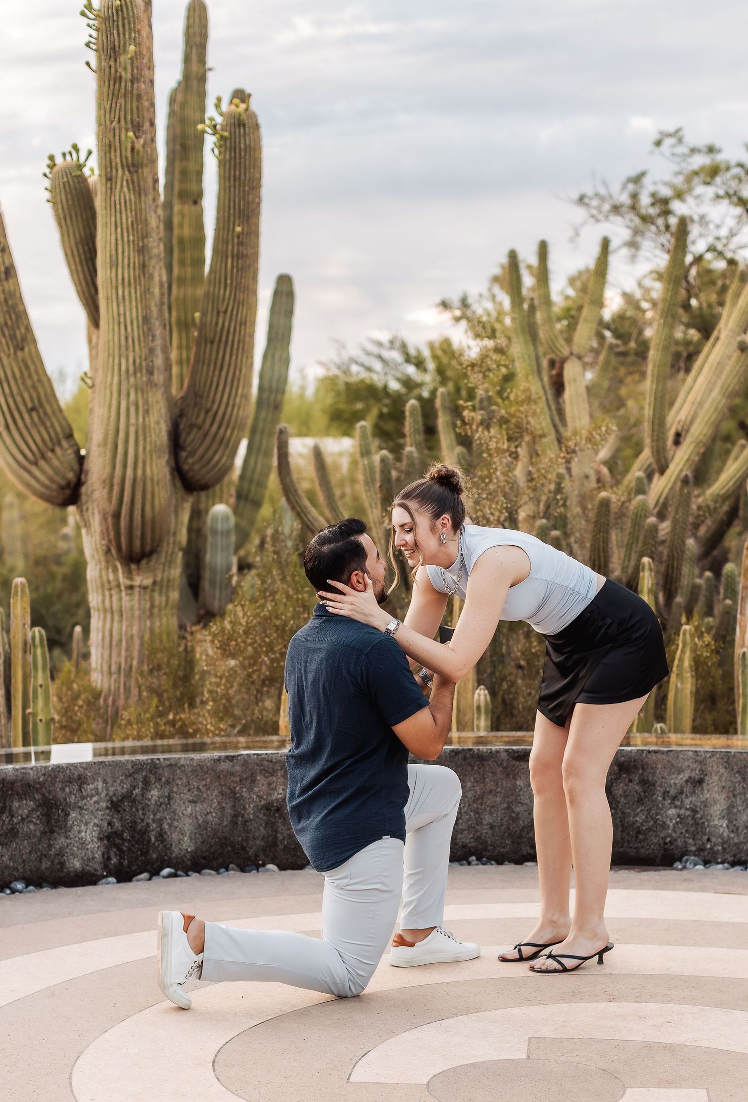 A romantic proposal moment captured in a desert garden surrounded by tall saguaro cacti on a cloudy day.