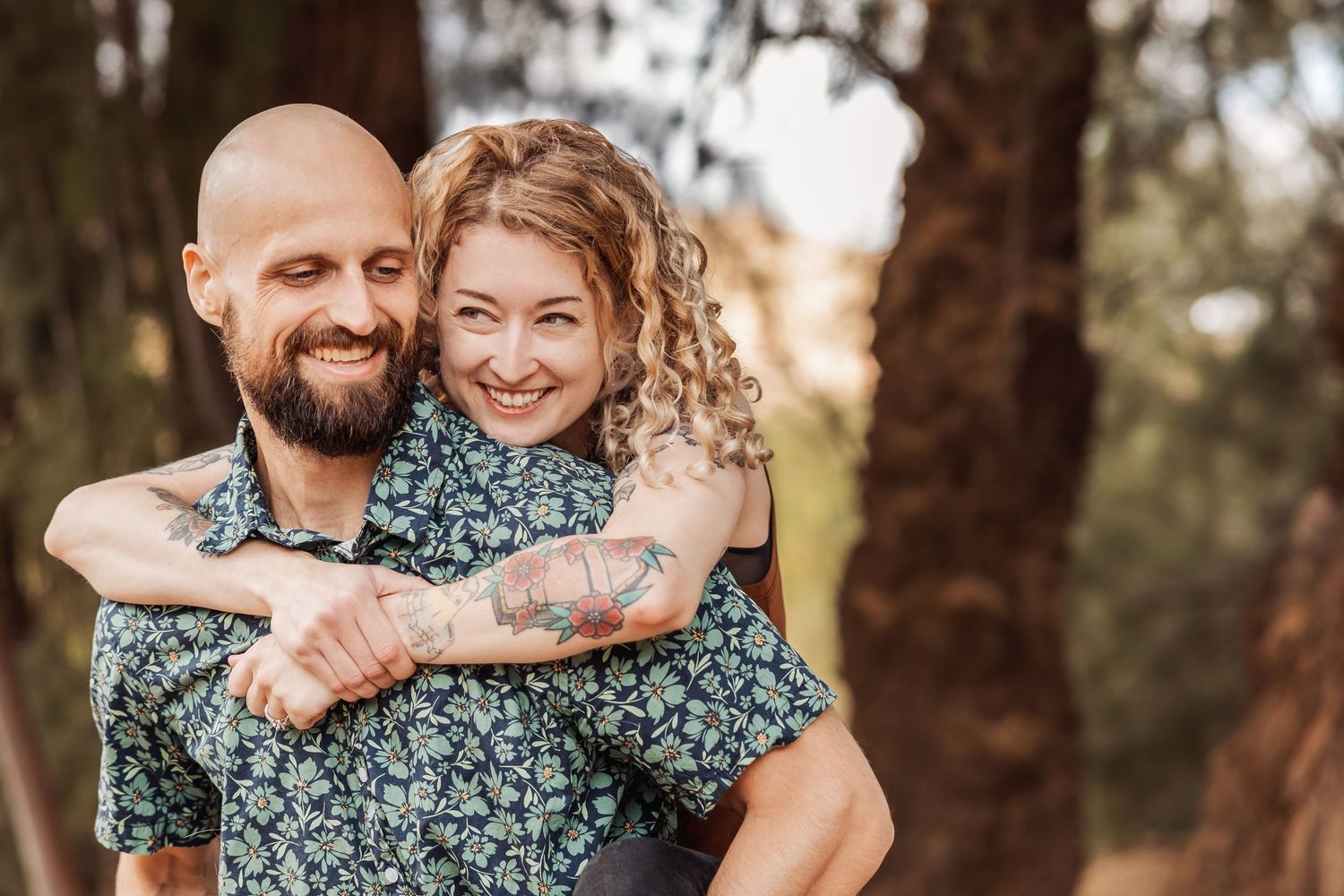 A couple wearing matching floral print shirts embrace in a warm sunlit forest setting during a photo session.
