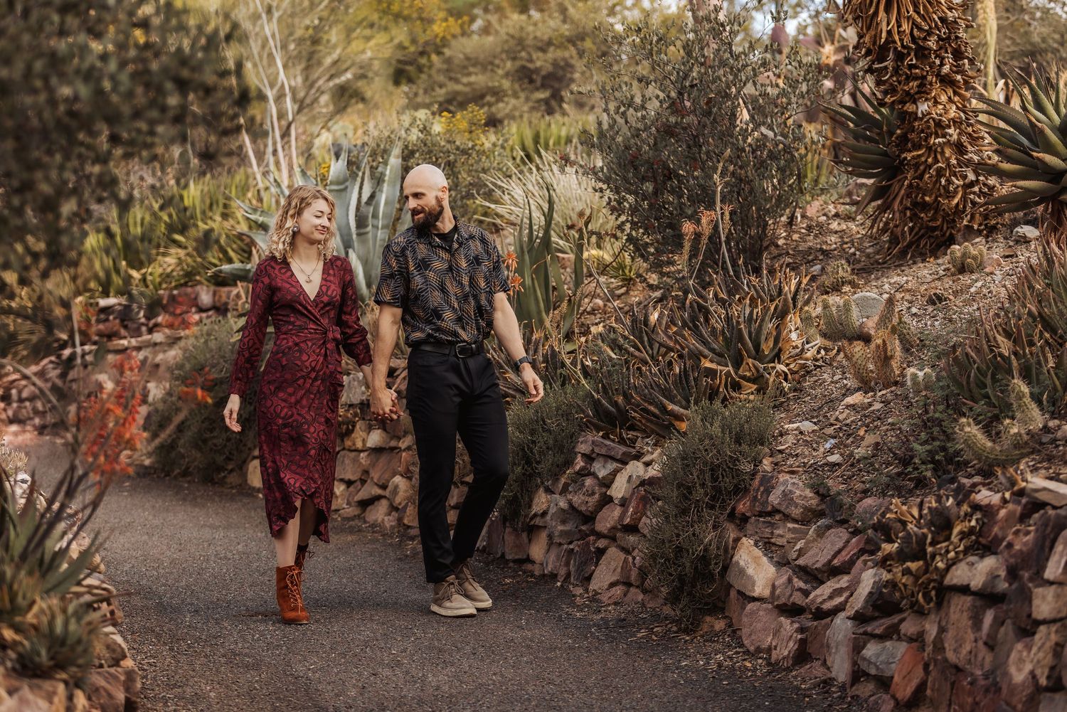 A couple walks hand in hand along a winding garden path surrounded by desert plants and rocky landscaping.