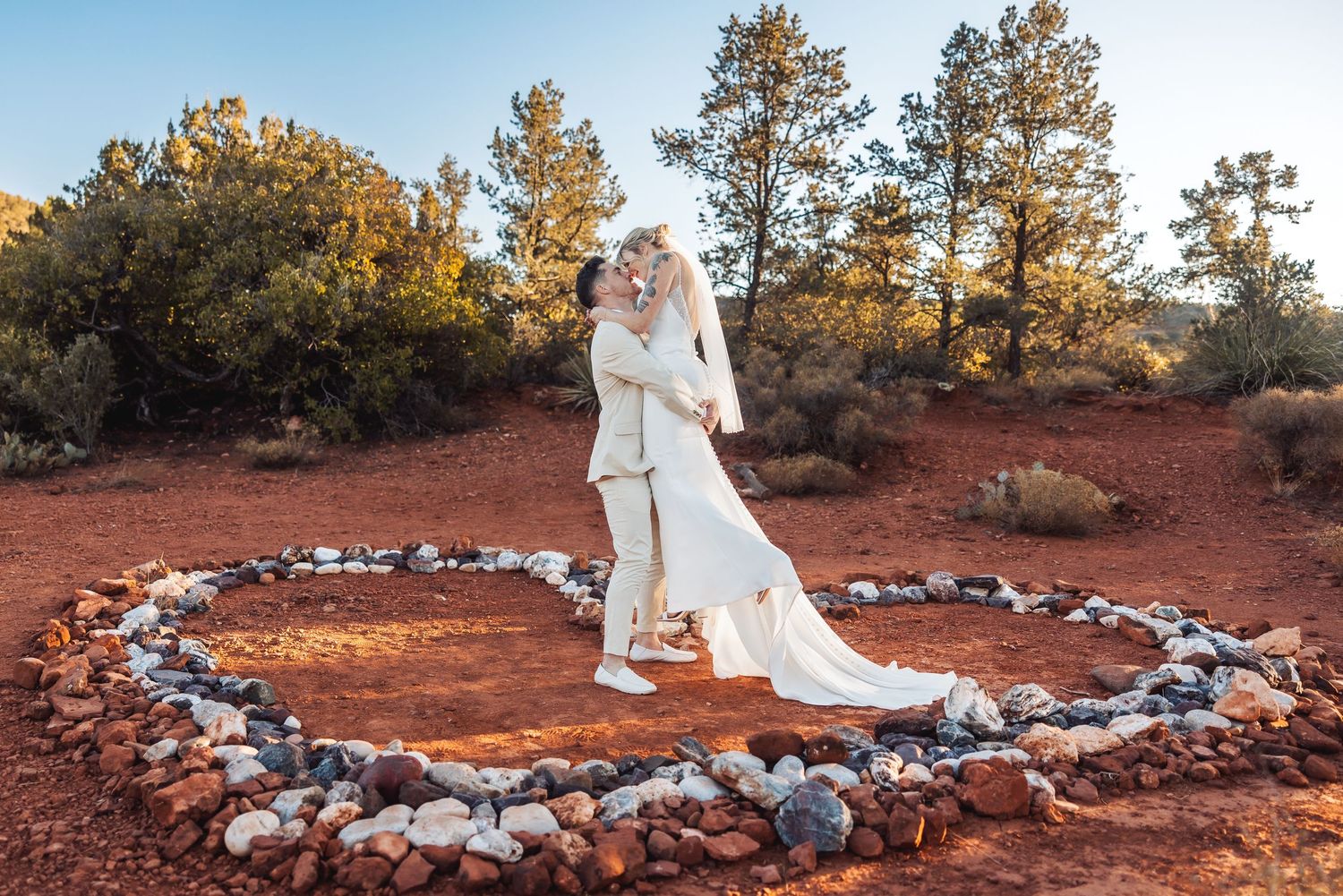 A wedding photo taken in red rocks desert landscape with heart-shaped stone arrangement and tall pine trees in background.