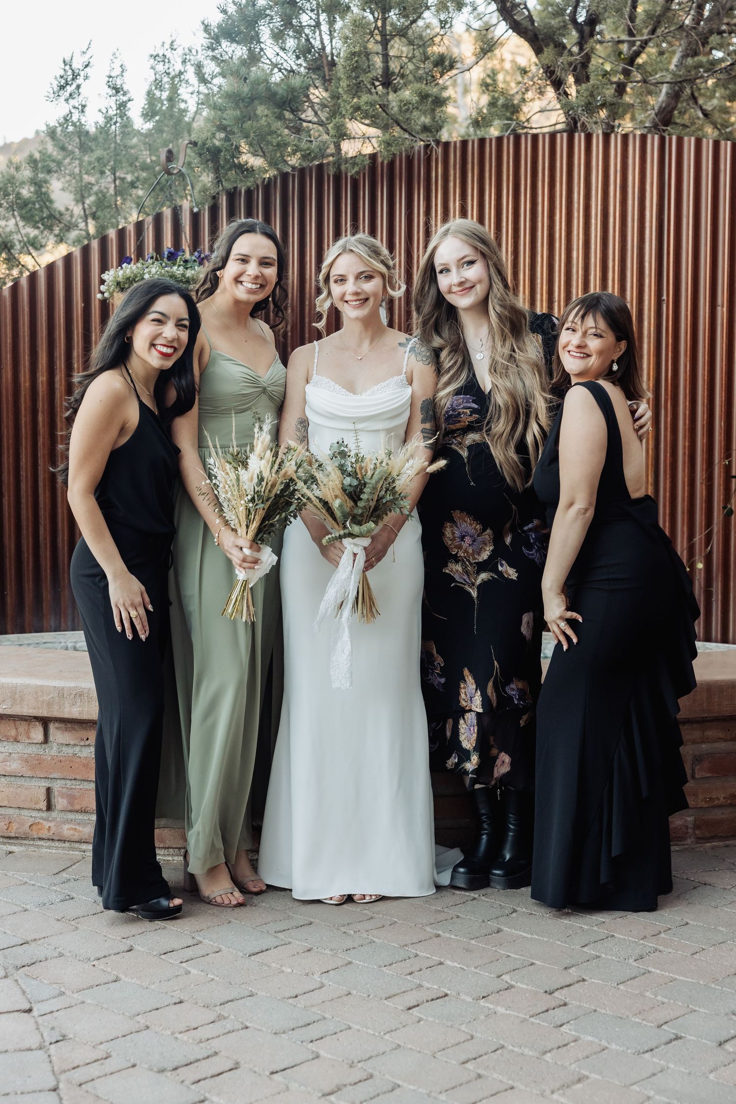 Bridal party poses together wearing elegant black and sage green dresses against wooden fence.