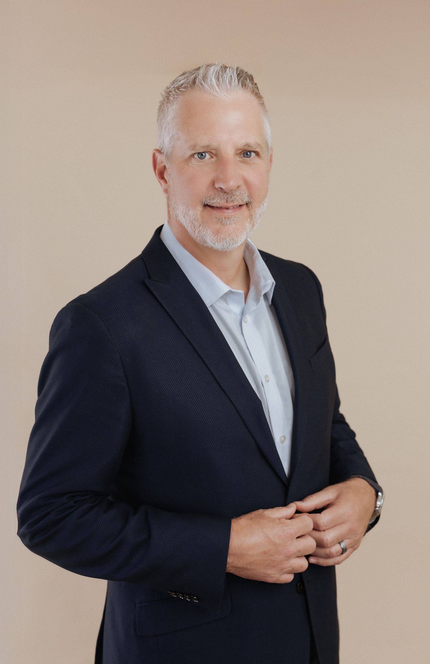 Professional headshot portrait of a person with gray hair and beard wearing a dark suit and light blue shirt.