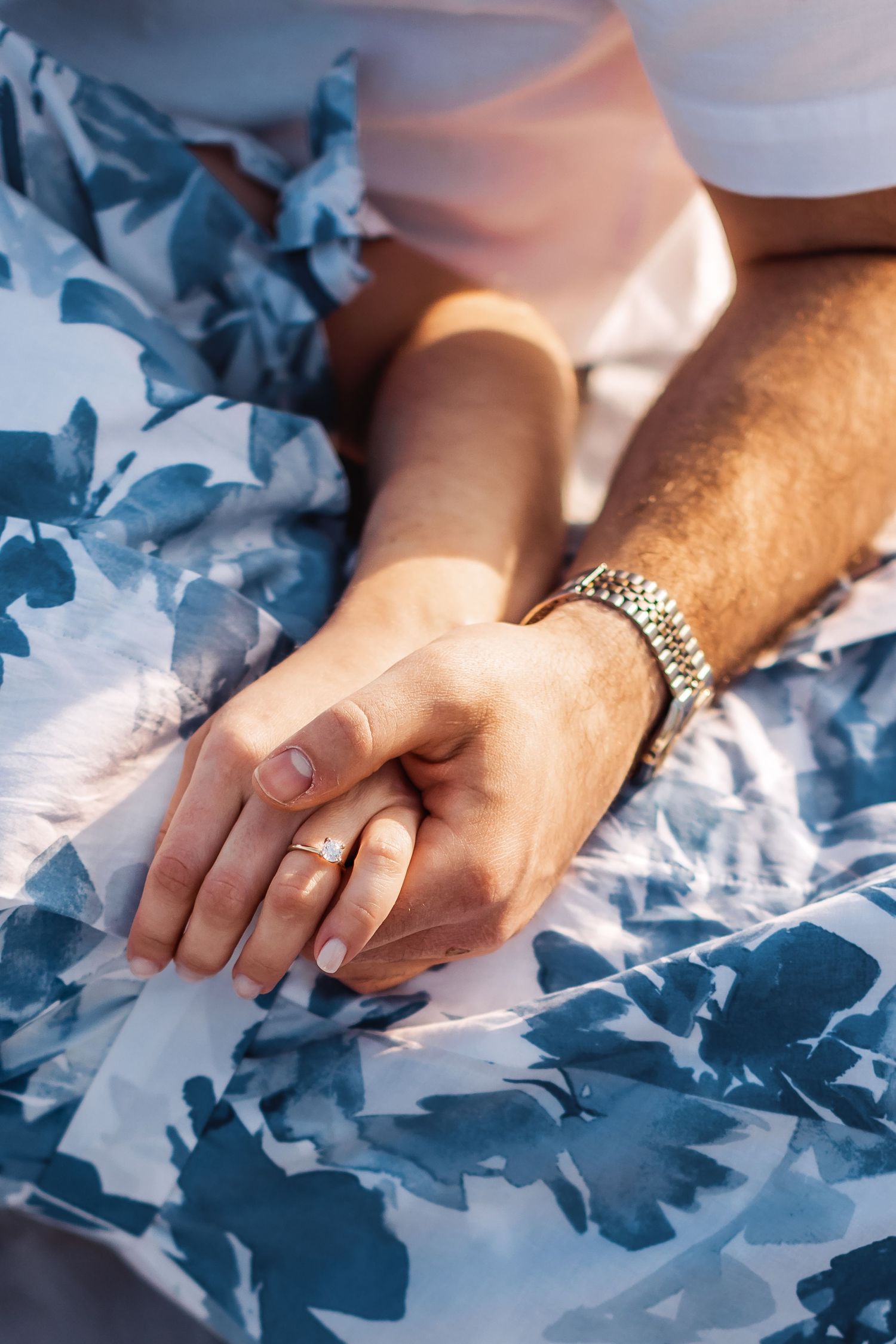 Two hands gently clasped together on blue floral fabric, one wearing a bracelet and wedding ring.