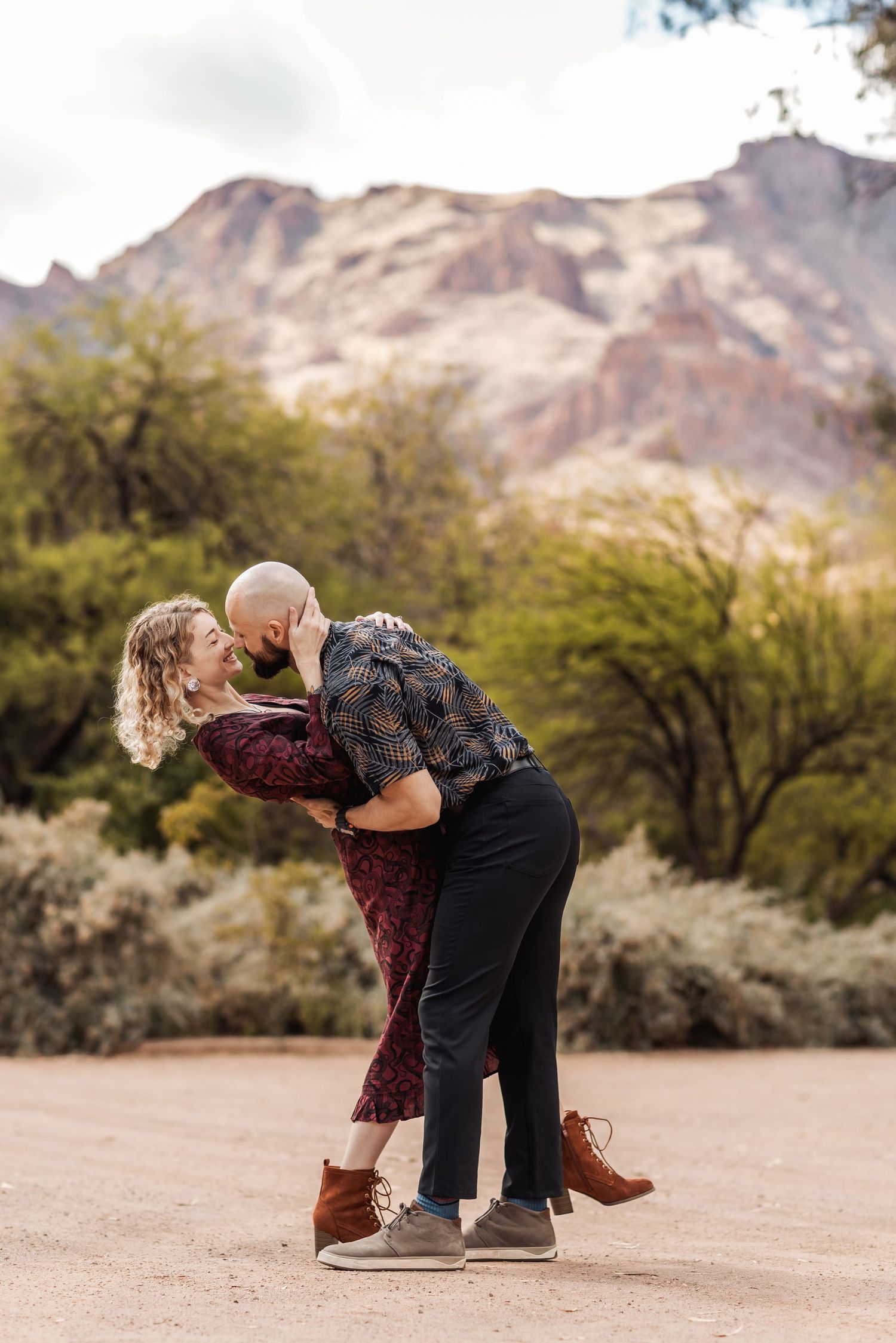 A romantic couple embraces and shares a kiss on a desert trail with rugged mountains in the background at sunset.