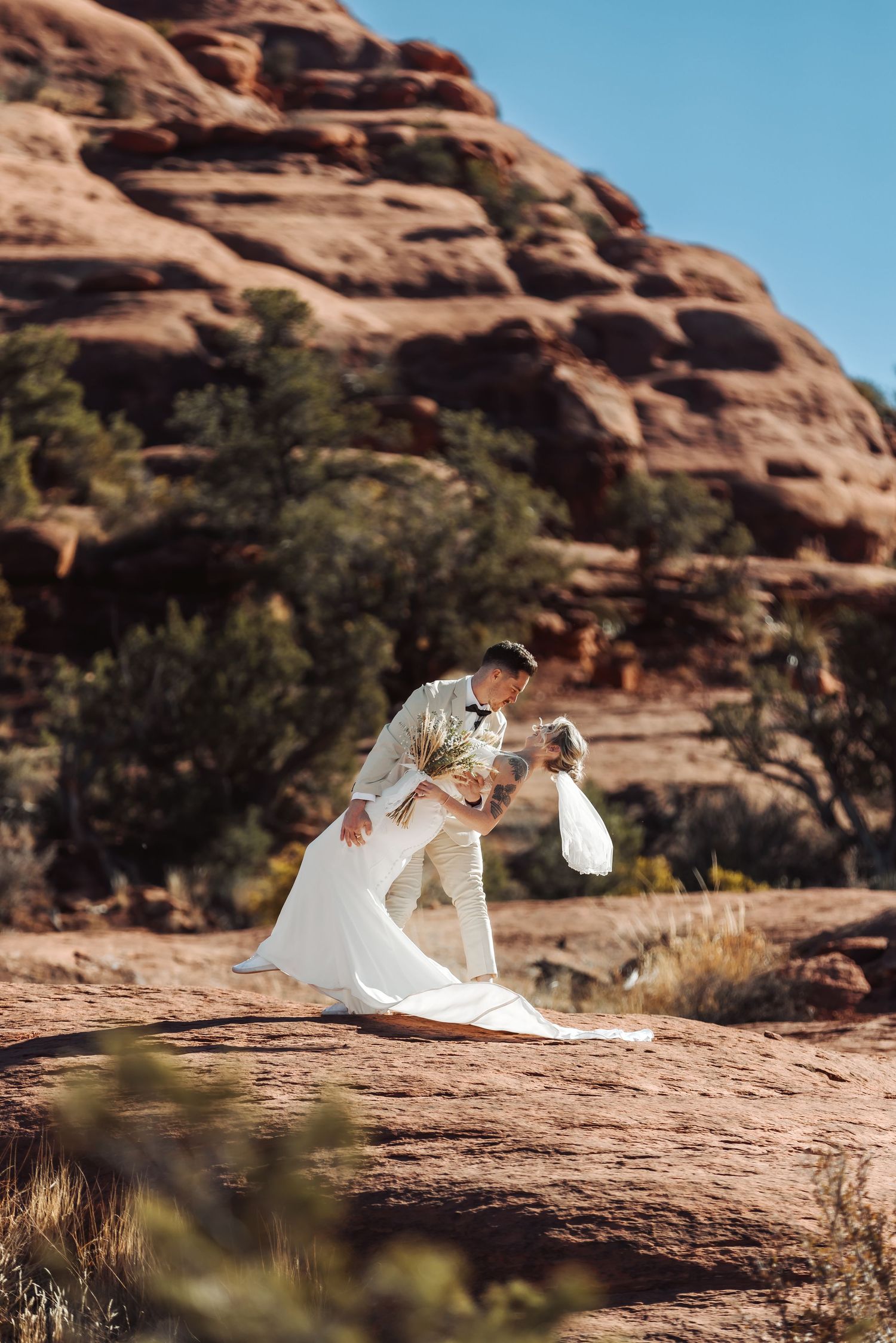 A romantic couple in white attire pose among the stunning red rock formations of a desert landscape.