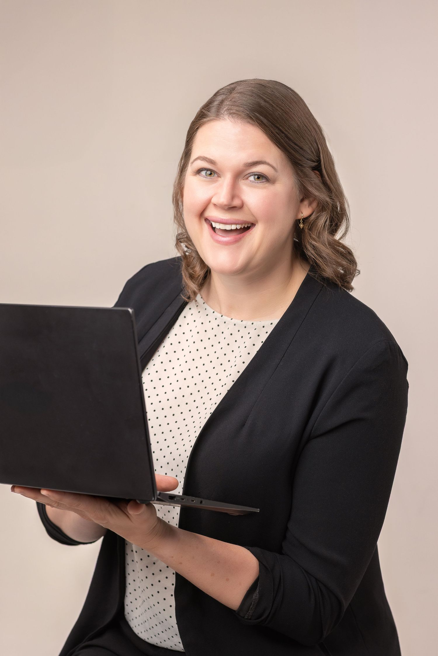 Professional wearing a black blazer and polka dot blouse holds a laptop while smiling warmly against neutral backdrop.