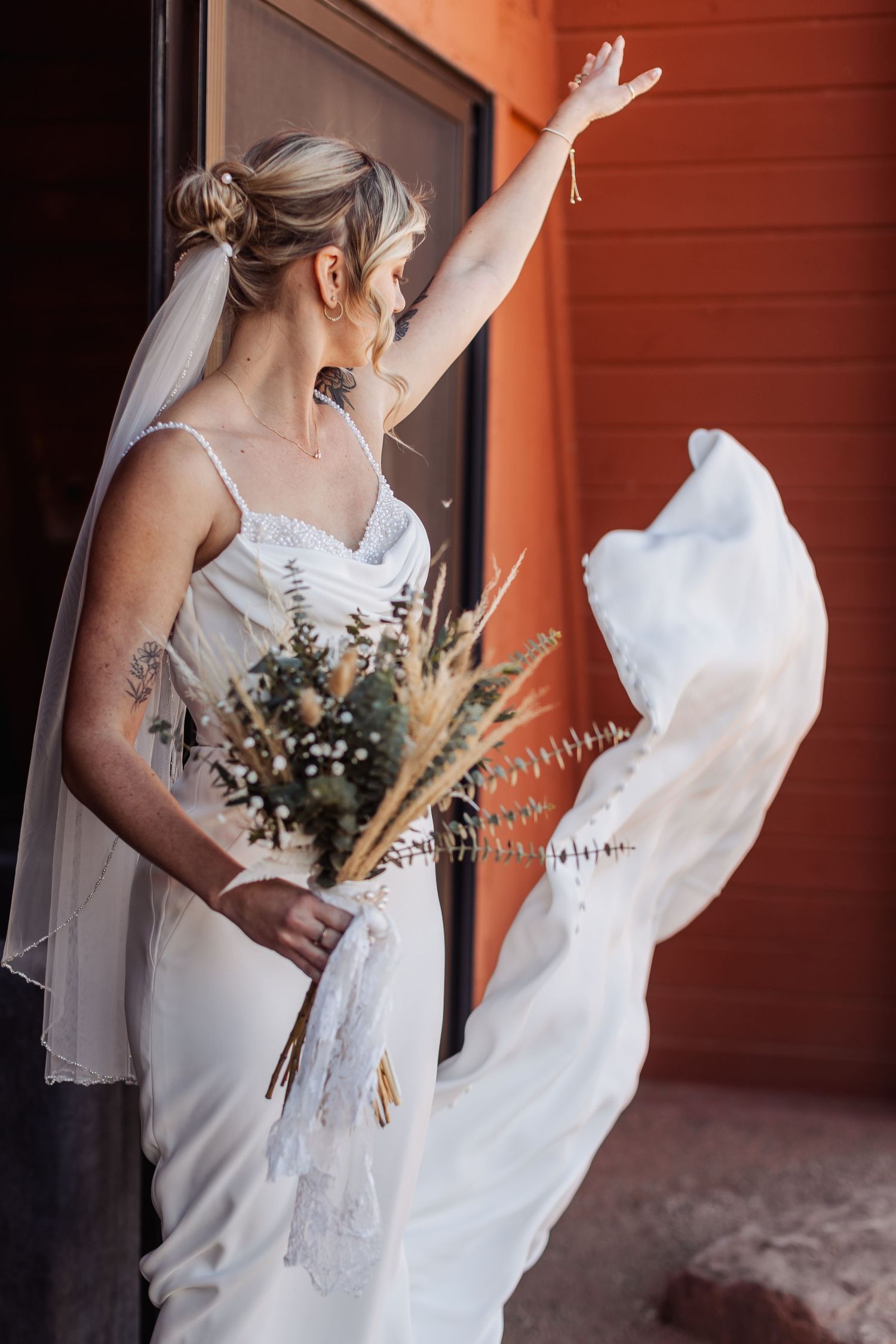 A bride in a white wedding dress holds a rustic bouquet with dried flowers and wheat stems against an orange wall.