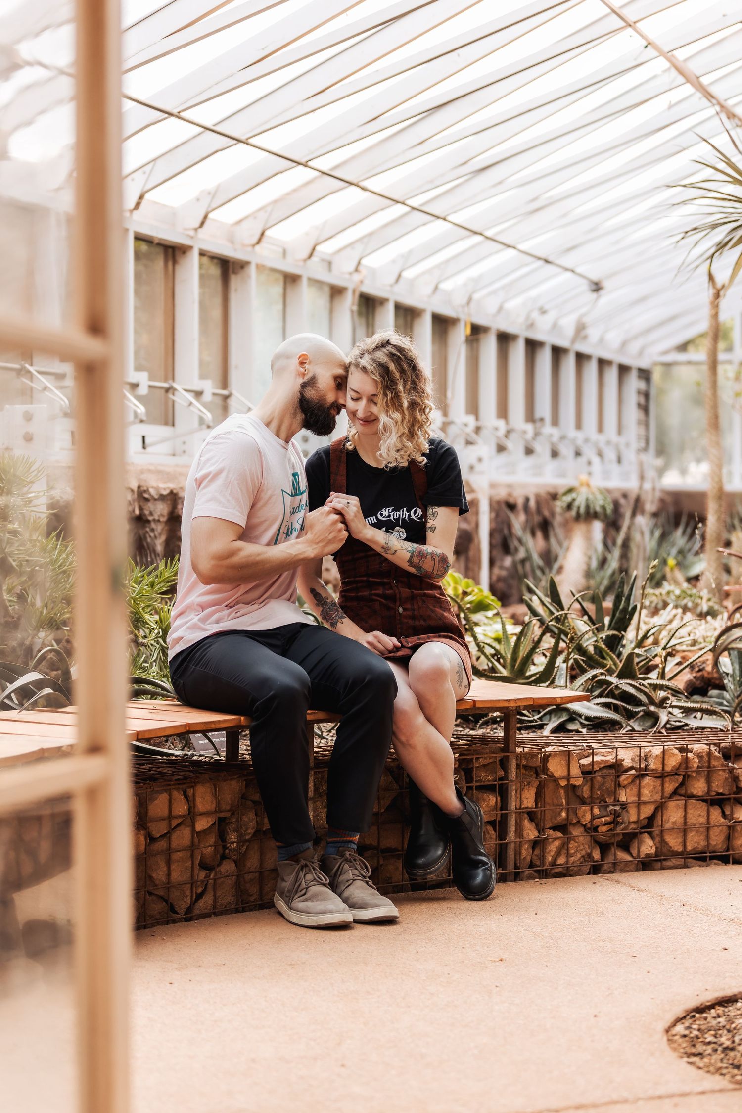 Two people share an intimate moment chatting while sitting together in a greenhouse with succulents and sunlit glass panels.
