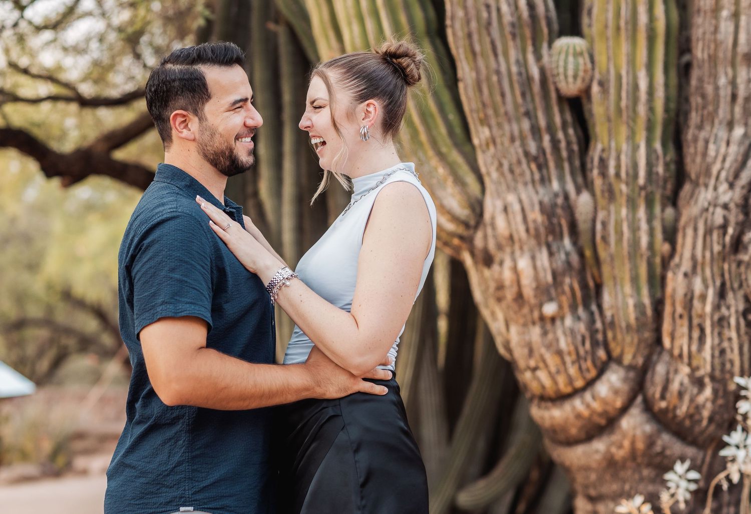 A couple embraces and smiles at each other while standing next to a large tree trunk in a natural outdoor setting.