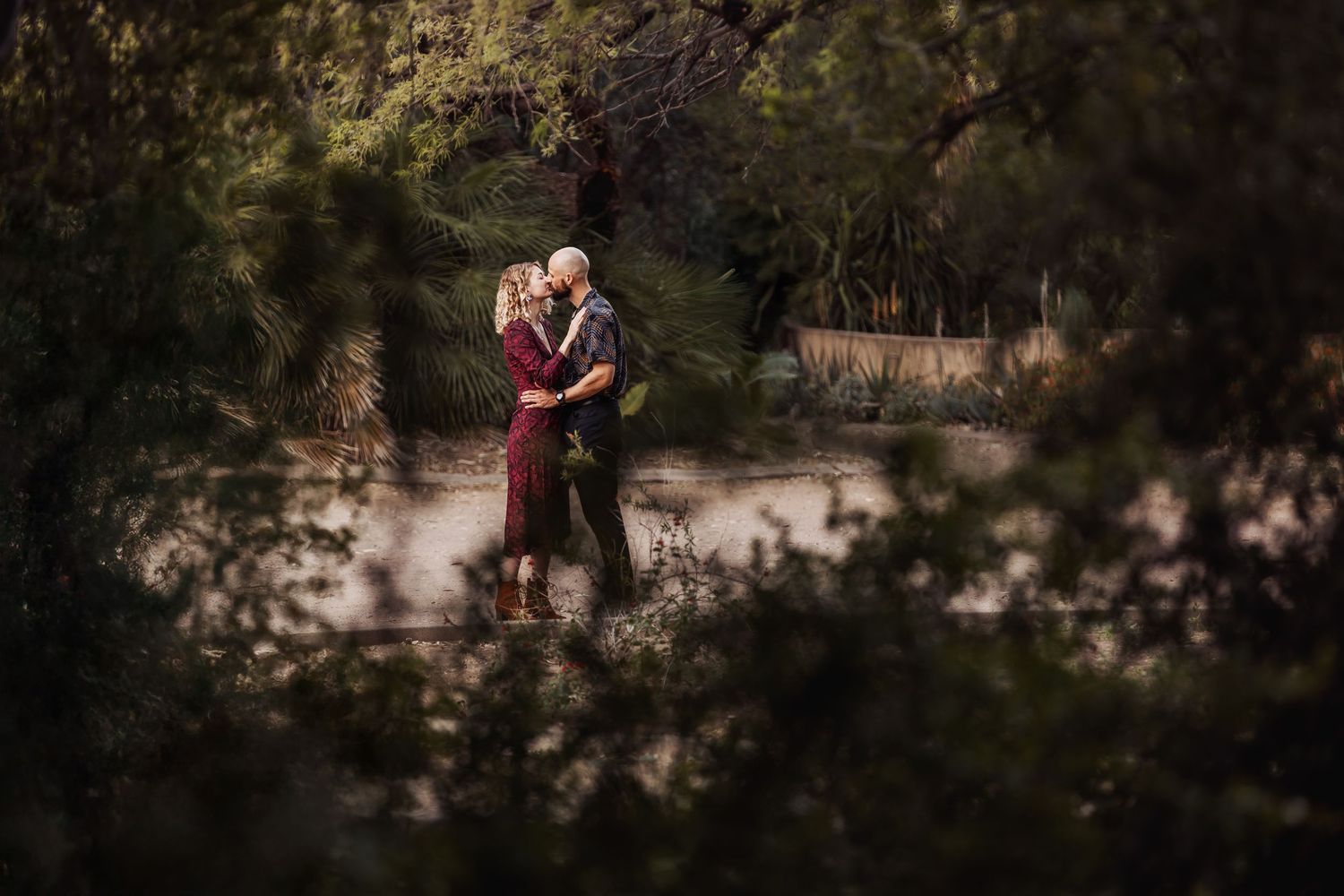 A couple embraces in burgundy attire against a dreamy forest backdrop with sunlit branches and foliage.