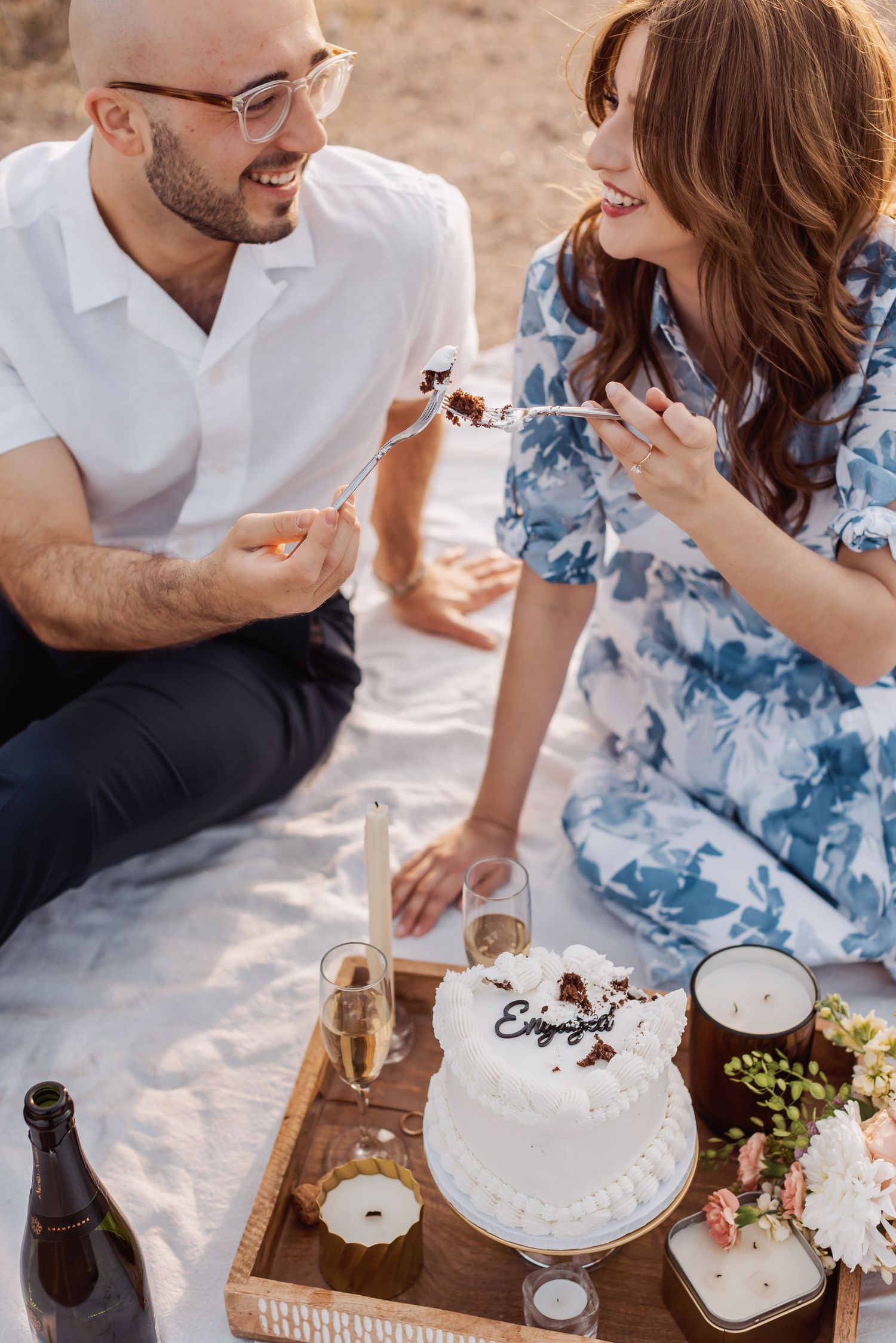 A couple enjoys a romantic picnic with wine, cake and grapes on a wooden tray in an outdoor setting.