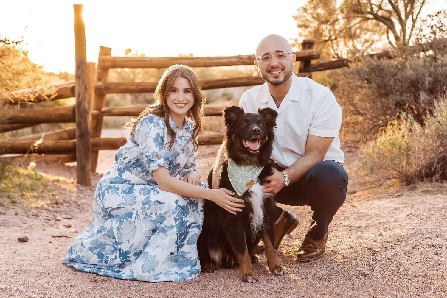 A couple kneels outdoors on a desert trail at sunset with their happy black dog in a rustic natural setting.