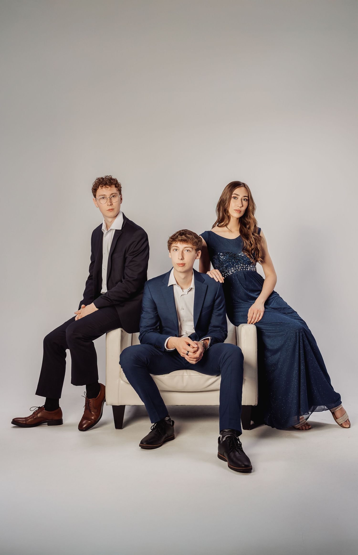 Three people in formal attire pose together on a light gray couch against a neutral background.