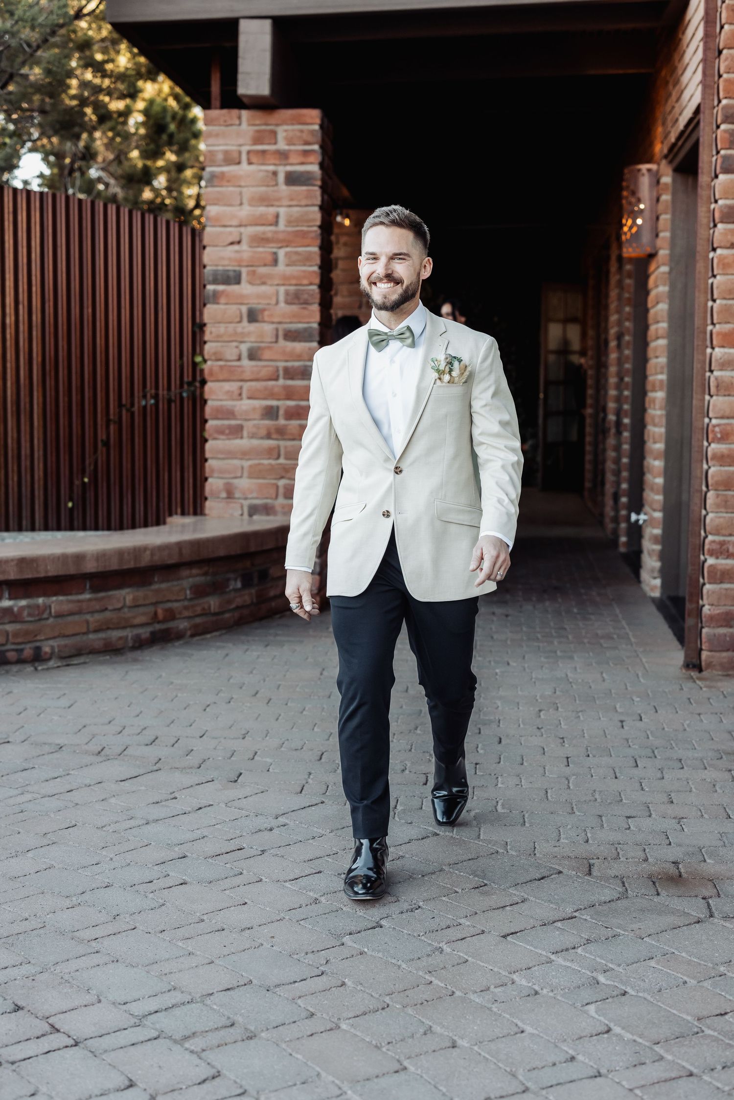 A groom in a cream-colored blazer and navy pants walks confidently on a brick-paved path during a wedding.