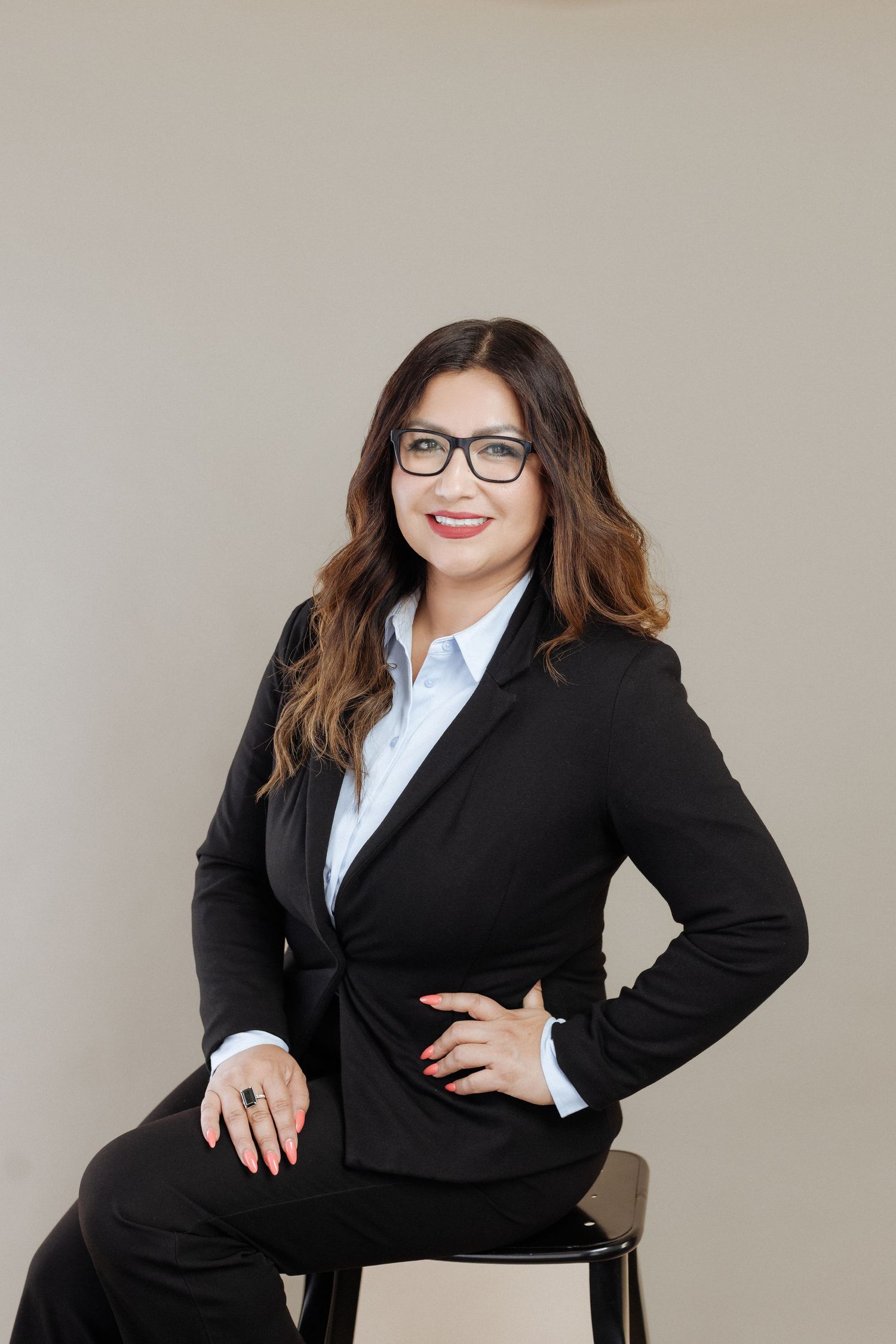Professional business photo of someone wearing a black suit and glasses sitting on a stool against a light background.
