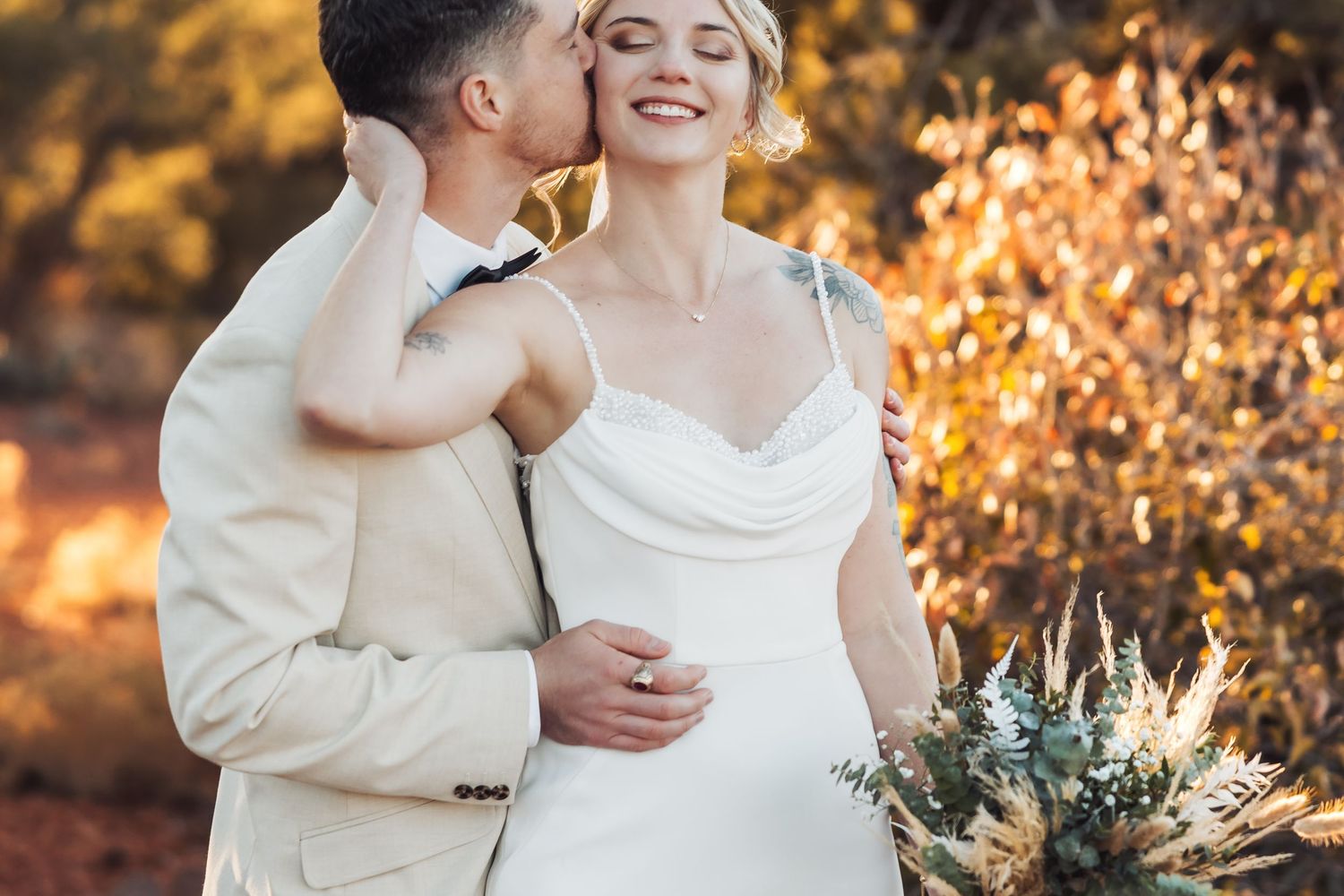 Romantic outdoor wedding portrait at sunset with golden autumn foliage and rustic floral bouquet.