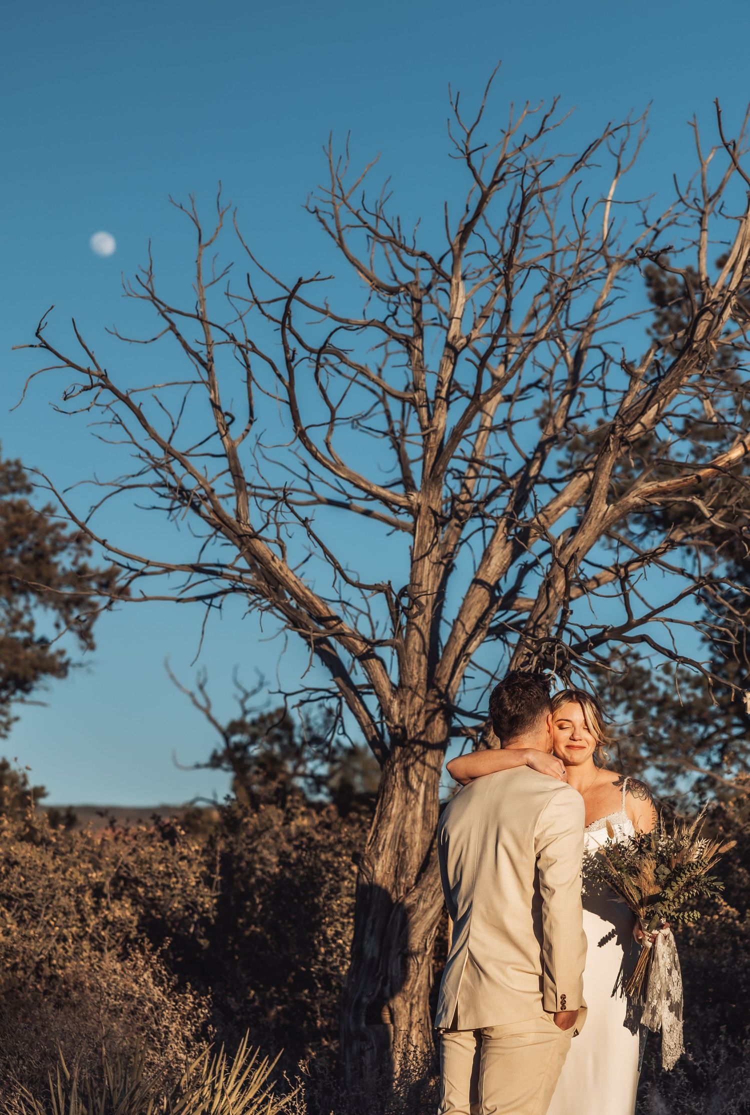 A romantic moment between a couple near a bare tree silhouetted against a bright blue sky with a visible moon.