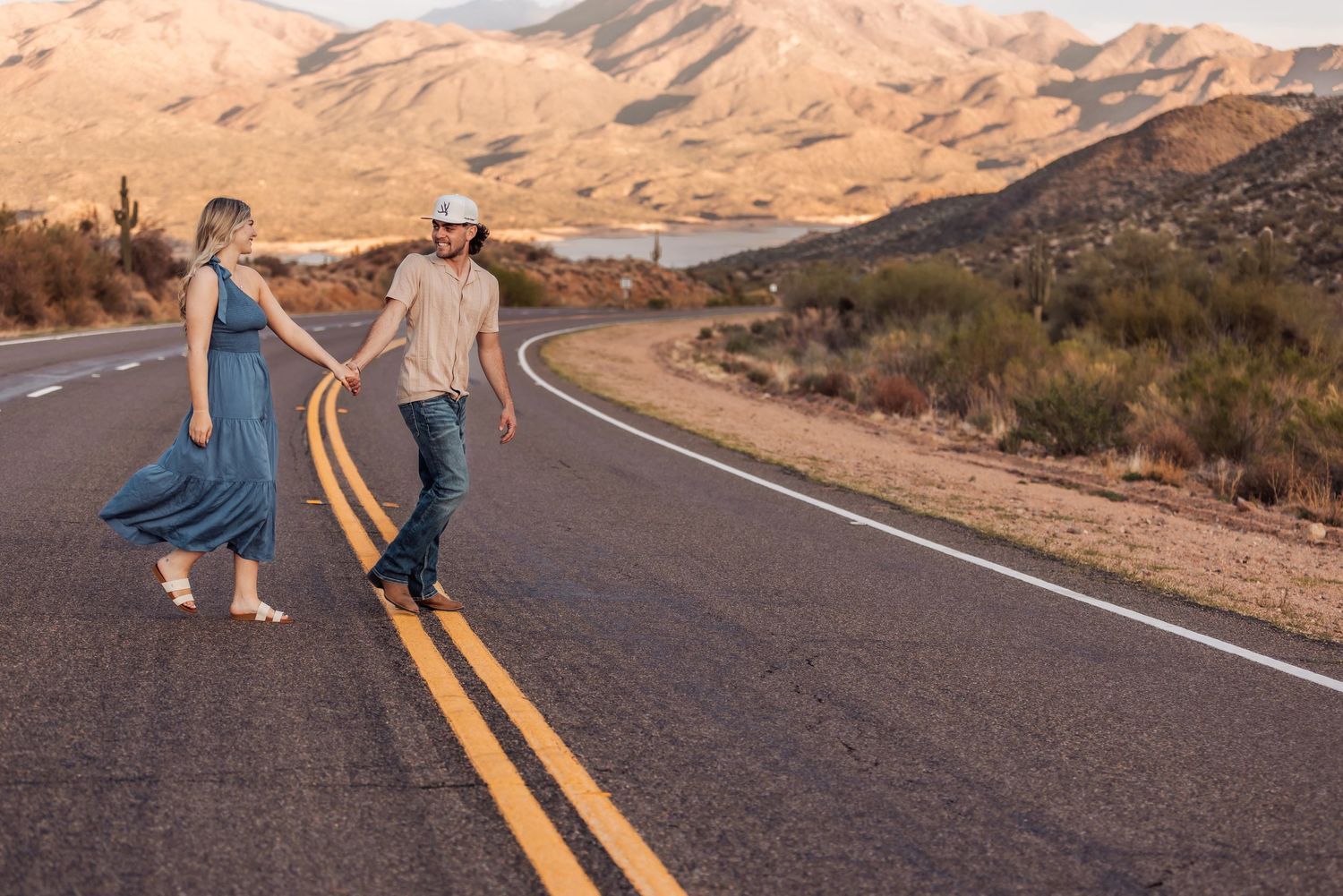 Two people in casual attire walk down the middle of an empty desert highway with mountains and cacti in the background.