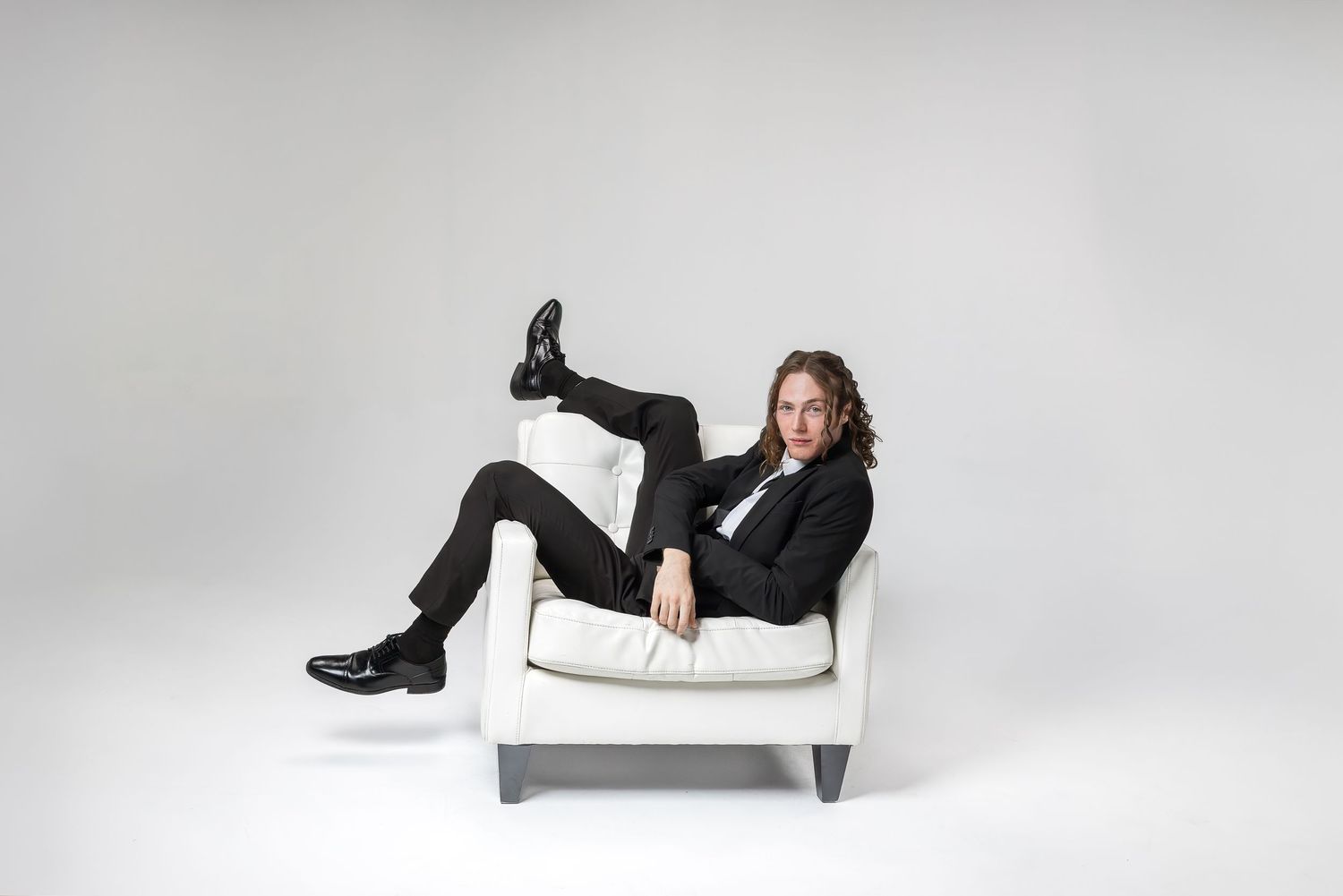 Person in black attire lounging playfully on white furniture against light studio backdrop.