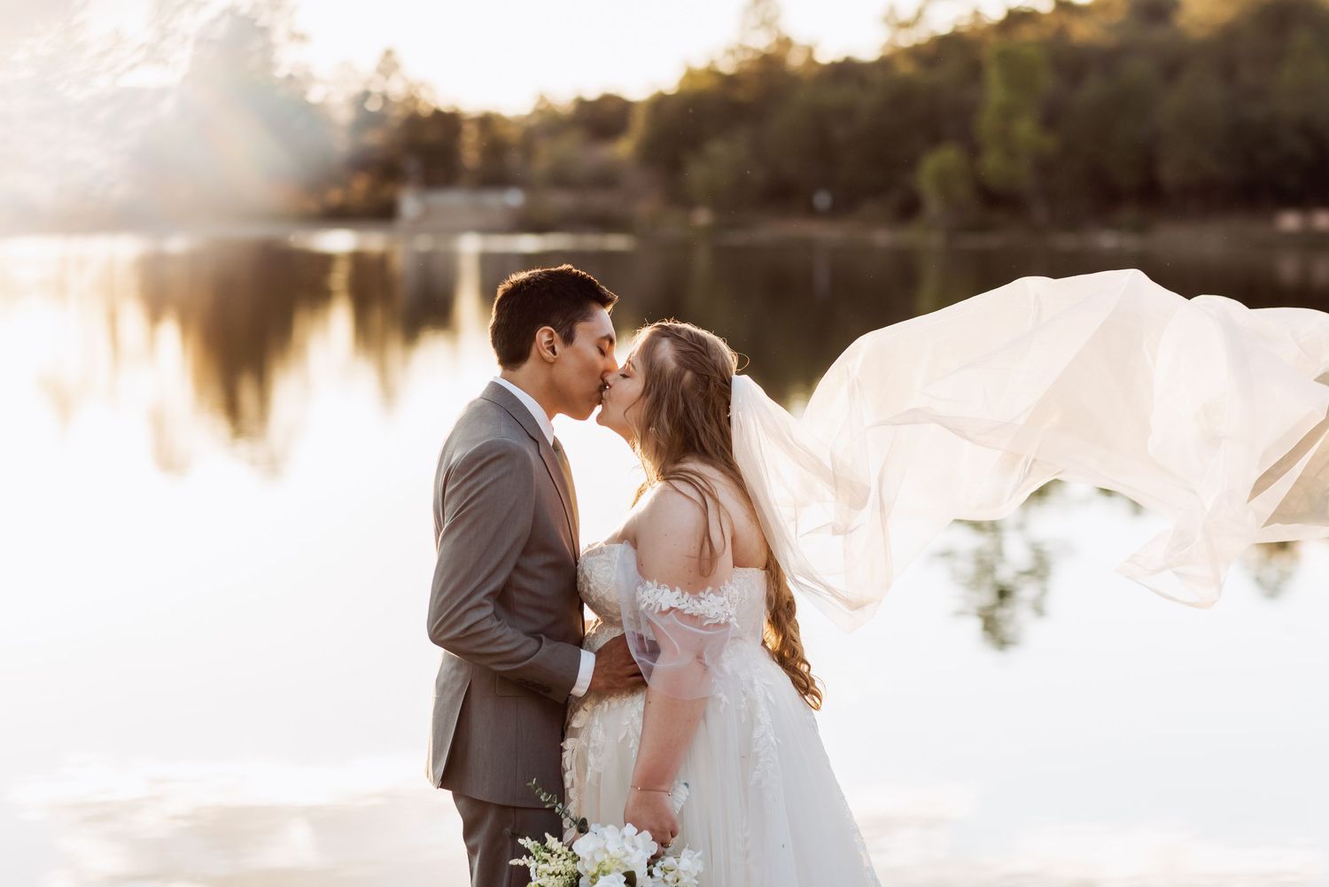 Romantic sunset wedding photo by a serene lake with bride in flowing white dress holding bouquet and groom in gray suit.