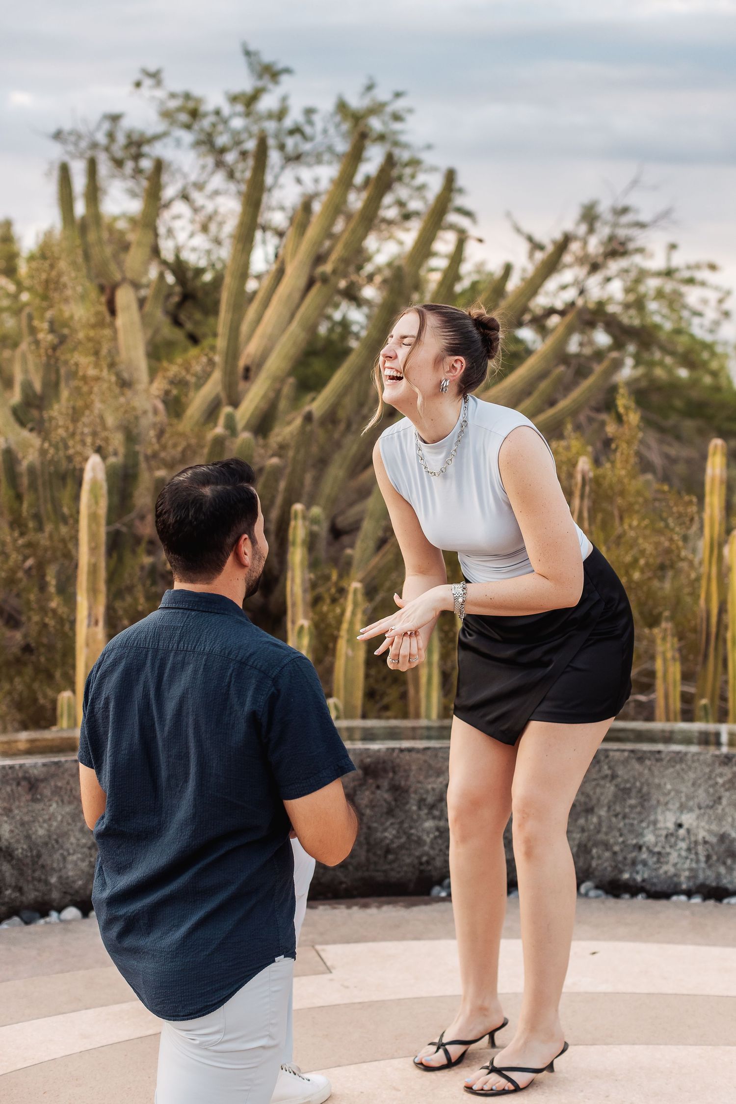 A couple sharing a joyful moment together in front of tall desert cacti during a sunset proposal.