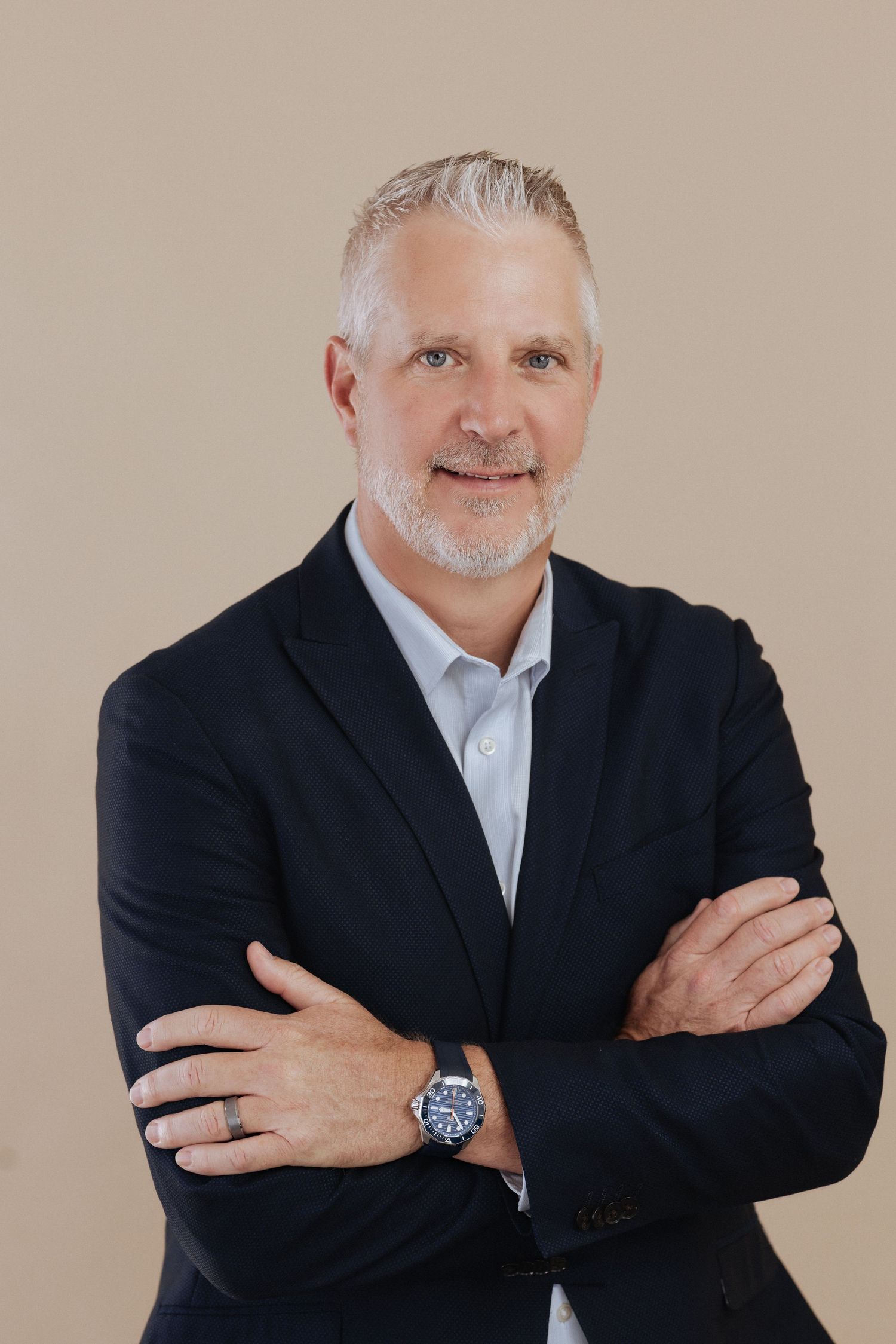 Professional business portrait of person with silver hair and beard wearing dark suit and white shirt against beige background.
