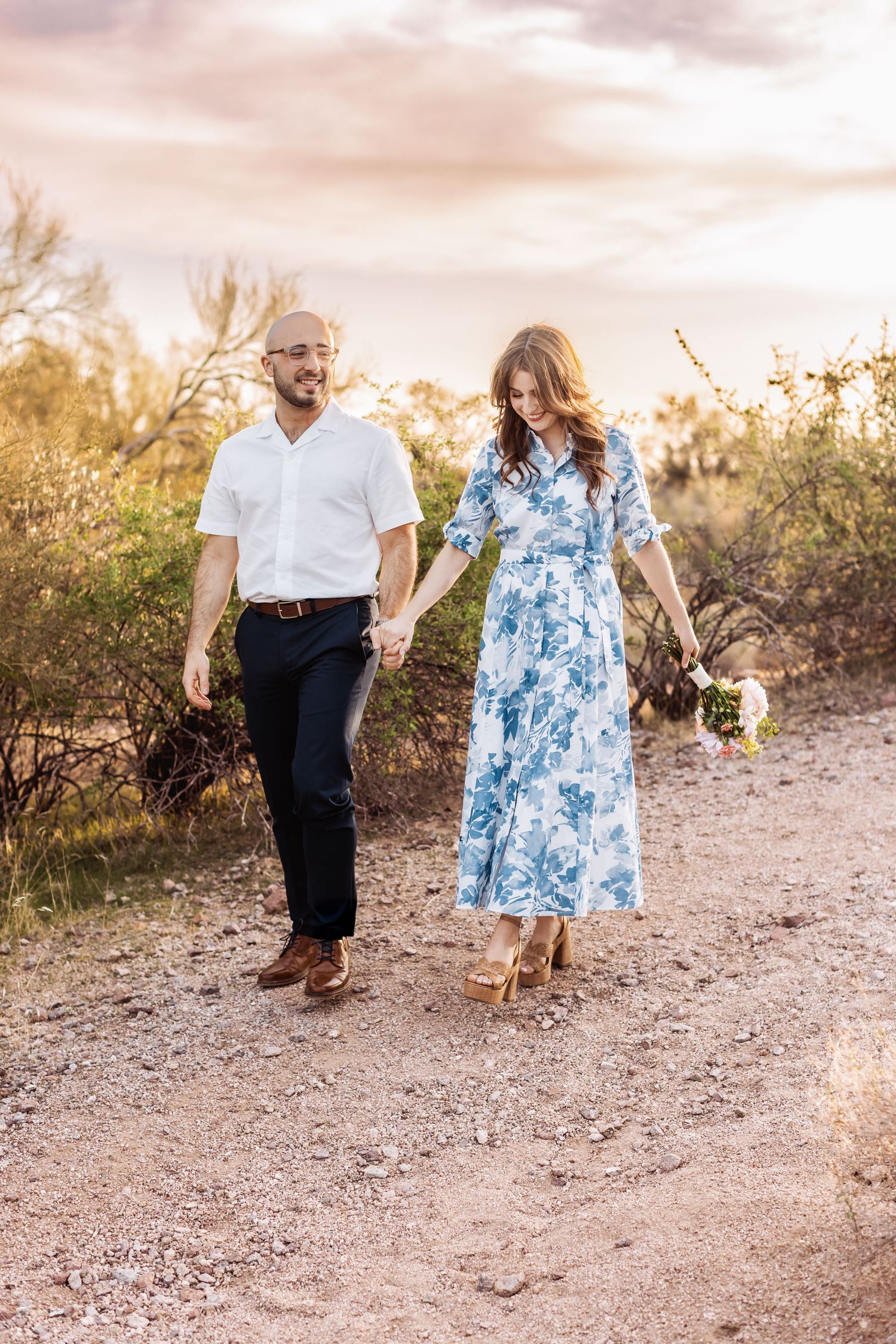 Two people walk hand-in-hand on a desert trail at sunset, with one wearing a blue floral dress and the other in a white shirt.