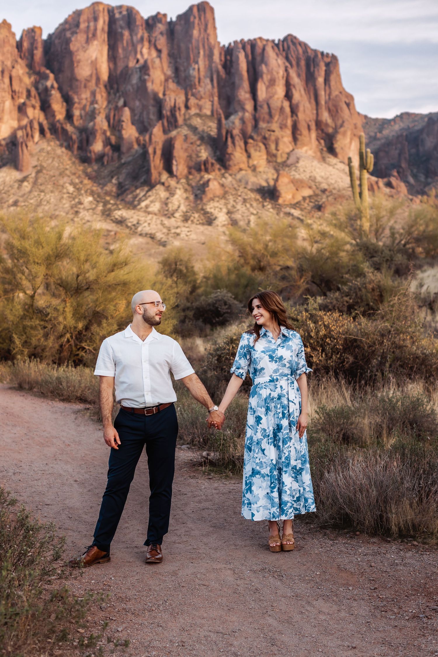 A couple walks hand in hand on a trail with the majestic Superstition Mountains and saguaro cacti behind them.