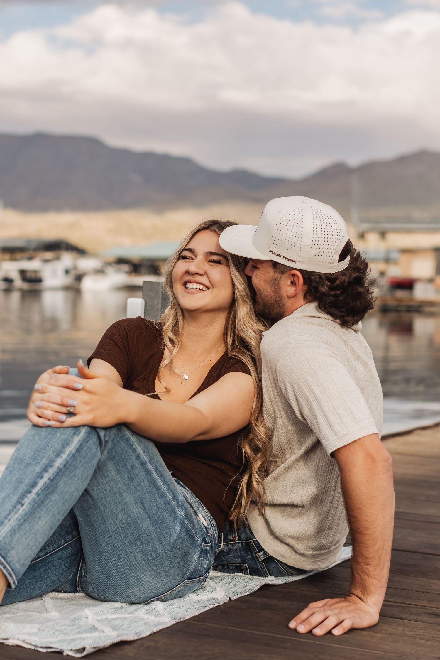 A couple sits together on a dock overlooking a marina with mountains in the background on a cloudy day.