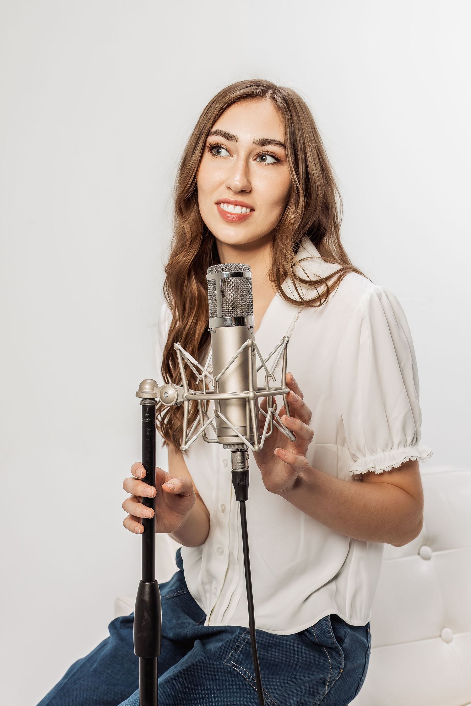 A woman in a white blouse sings into a professional studio microphone against a light background.