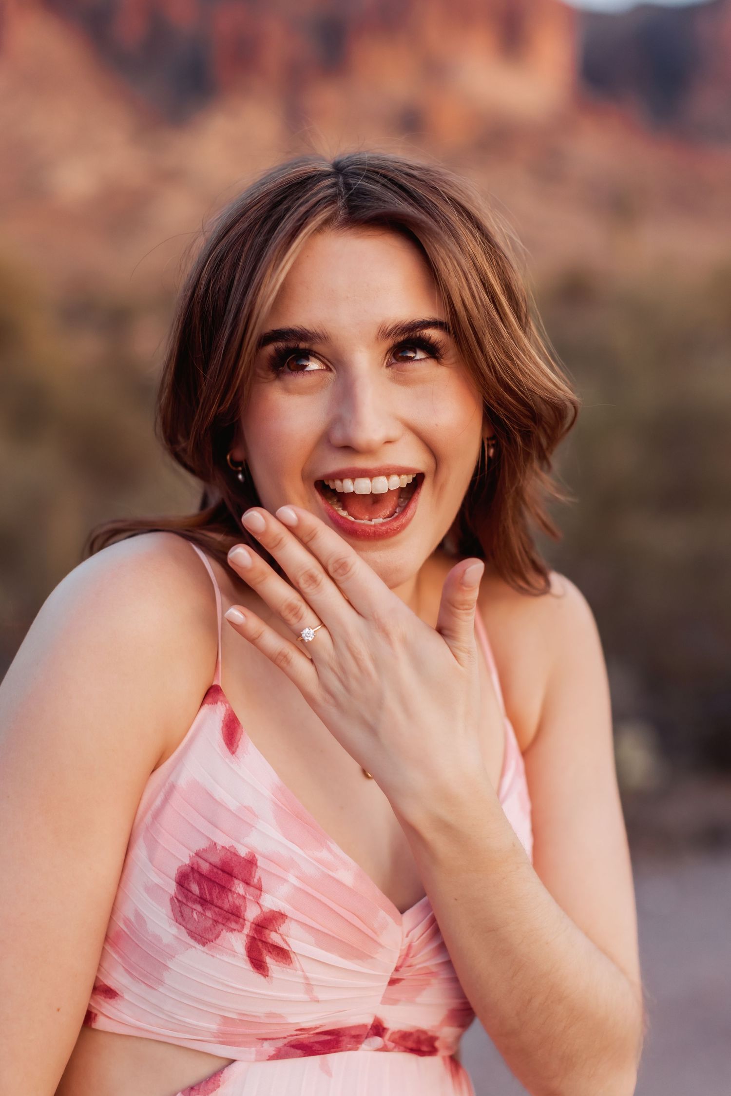 Woman in pink dress shows excitement while displaying ring against desert mountain backdrop.