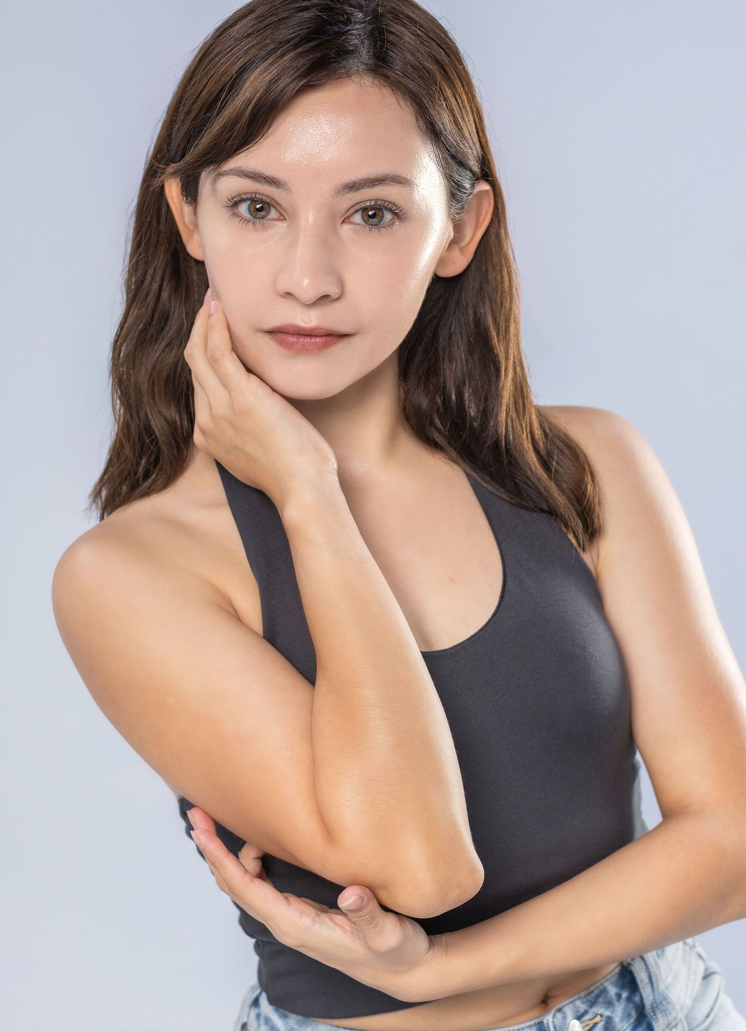 Young woman in black athletic tank top and casual attire poses with hand on face against light background.