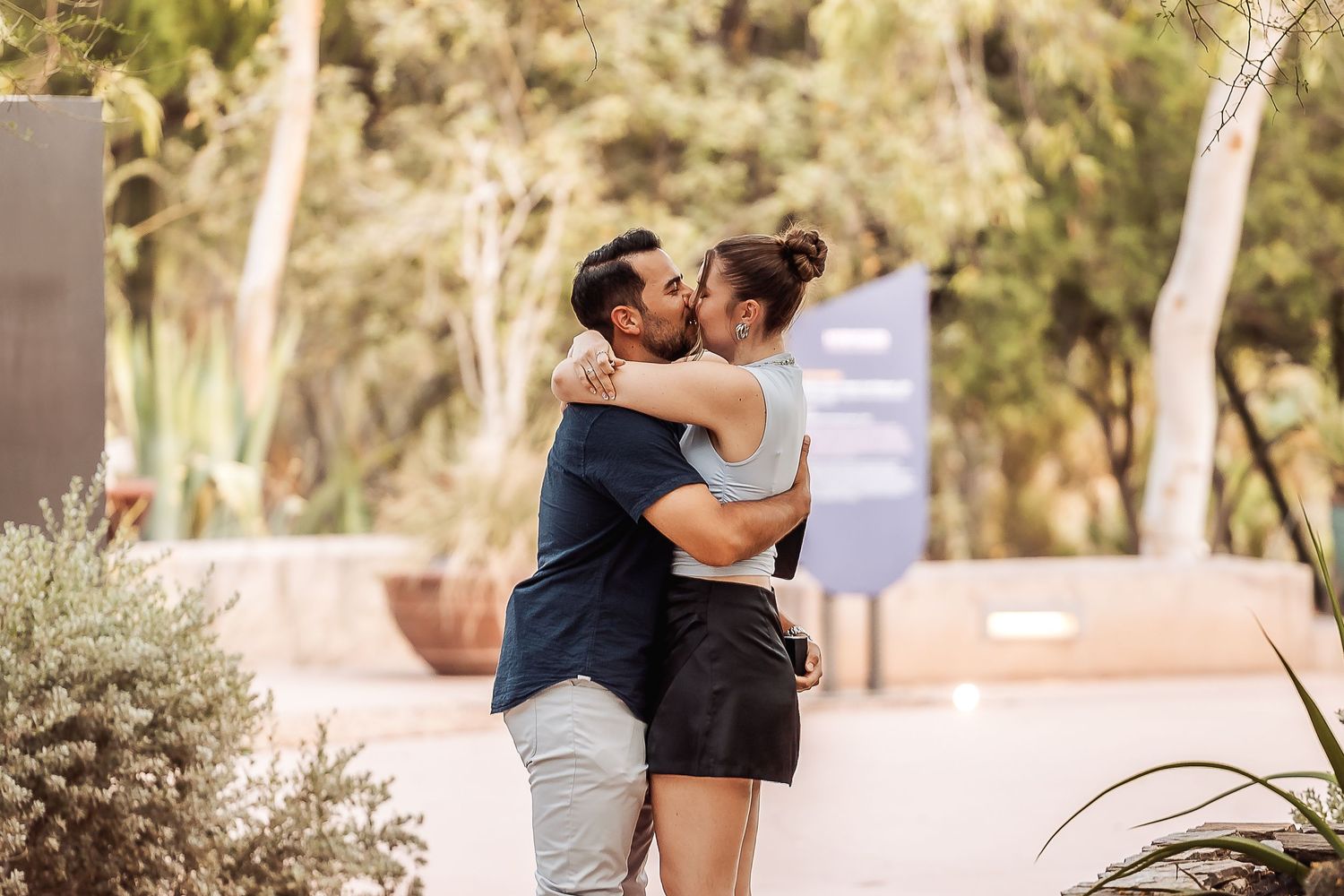 A couple embraces intimately in a sunlit garden pathway surrounded by trees and greenery.
