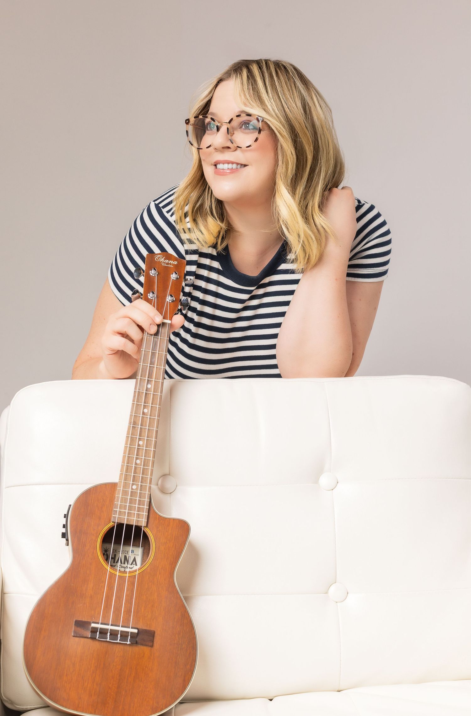 A person in a striped shirt lounges on a white couch with a ukulele, wearing glasses and smiling.
