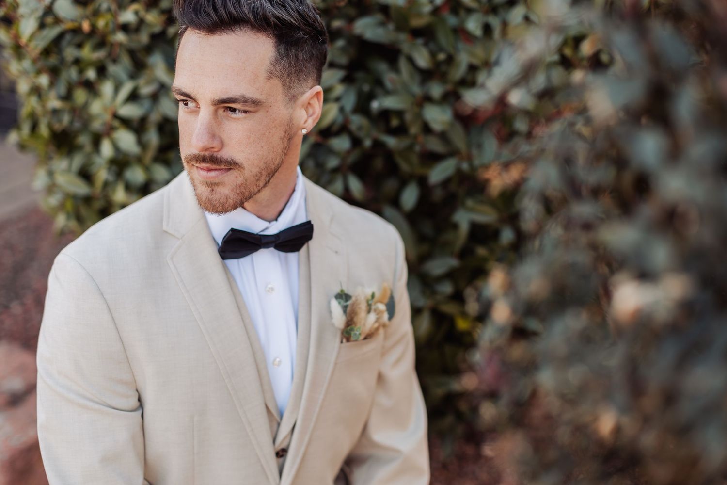 Man in beige suit jacket and bow tie poses outdoors against greenery.