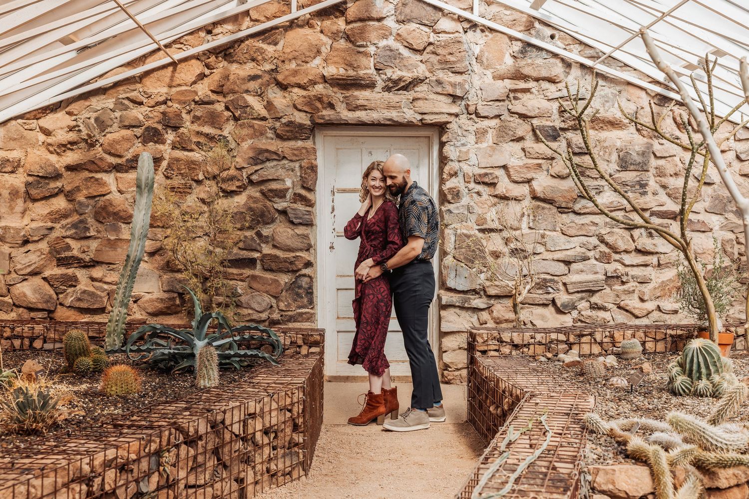 Couple embraces in doorway of rustic stone greenhouse with potted cacti and desert plants lining walkway.