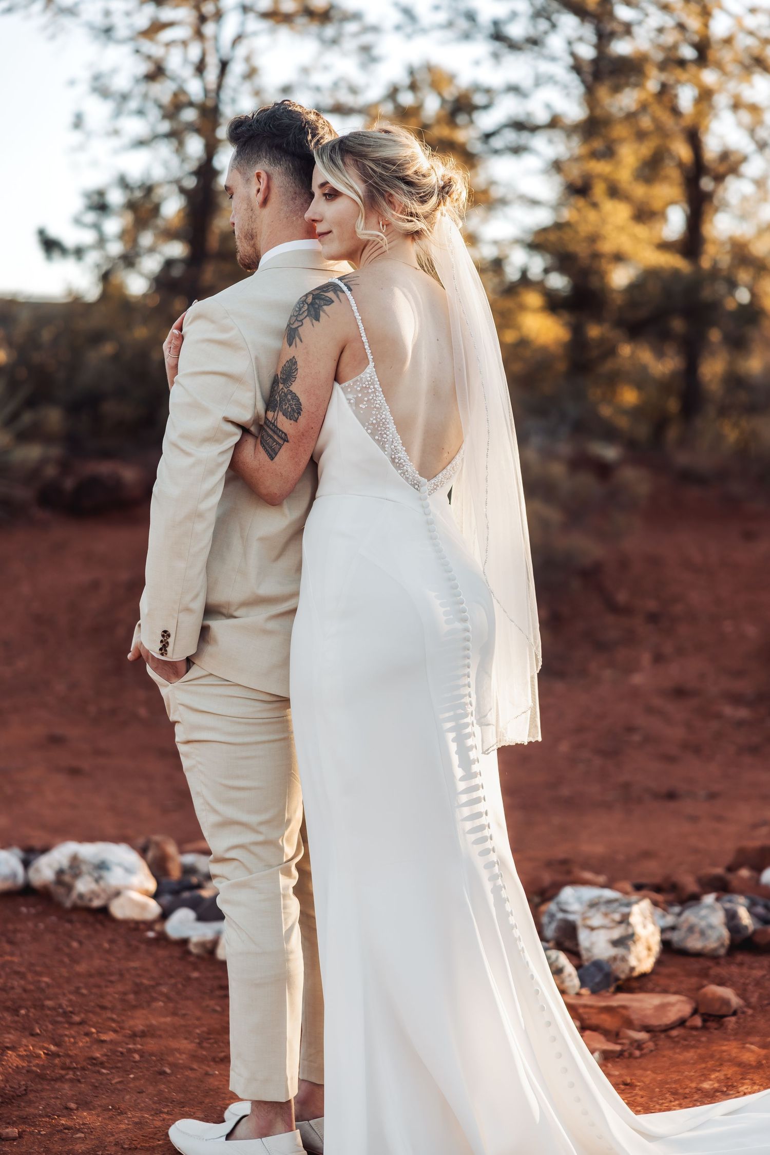 A couple in wedding attire embraces on red desert soil at sunset, wearing cream and white formal wear with a flowing veil.