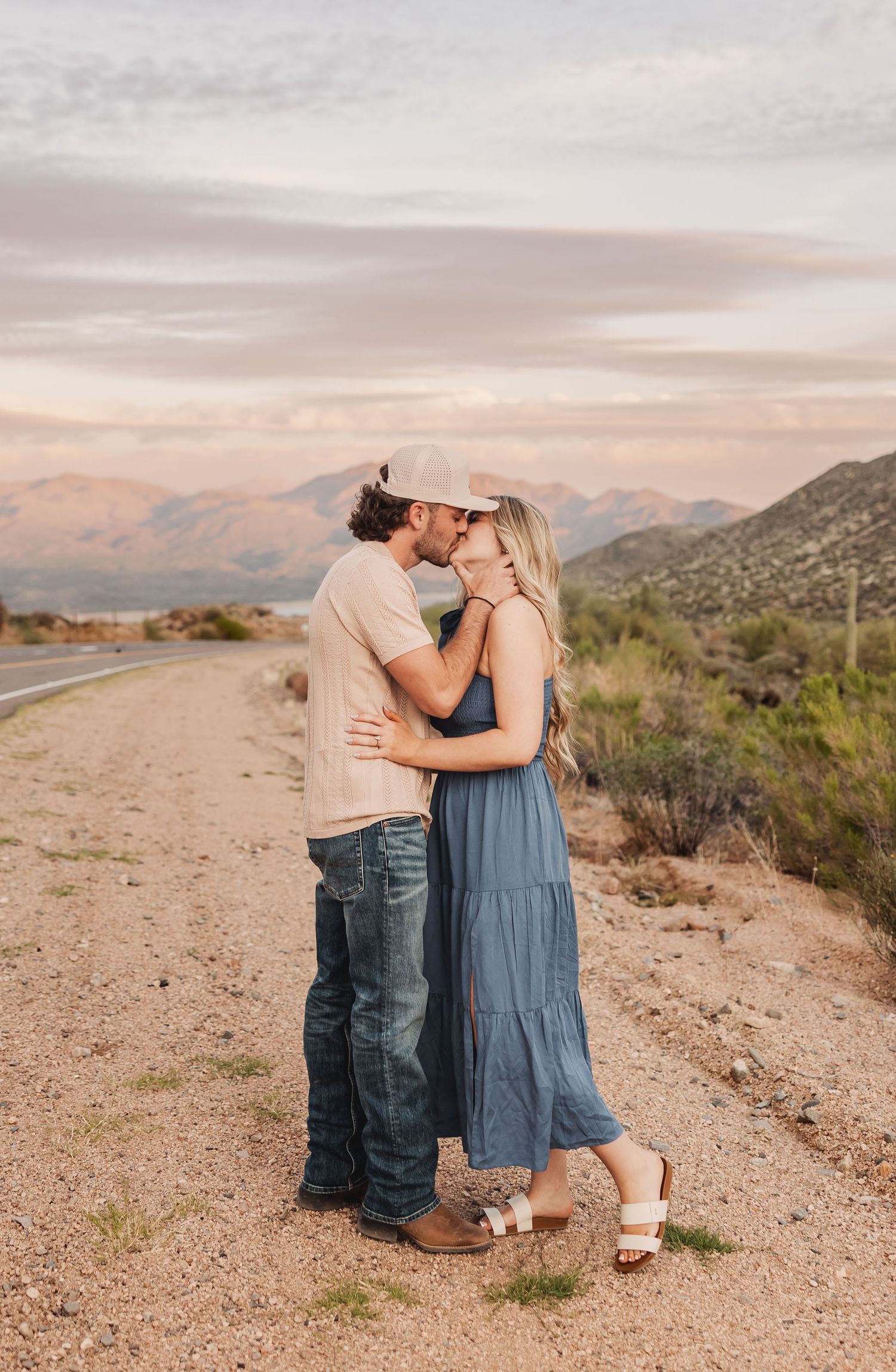 A romantic couple embraces on a desert road at sunset with mountains and desert landscape in the background.