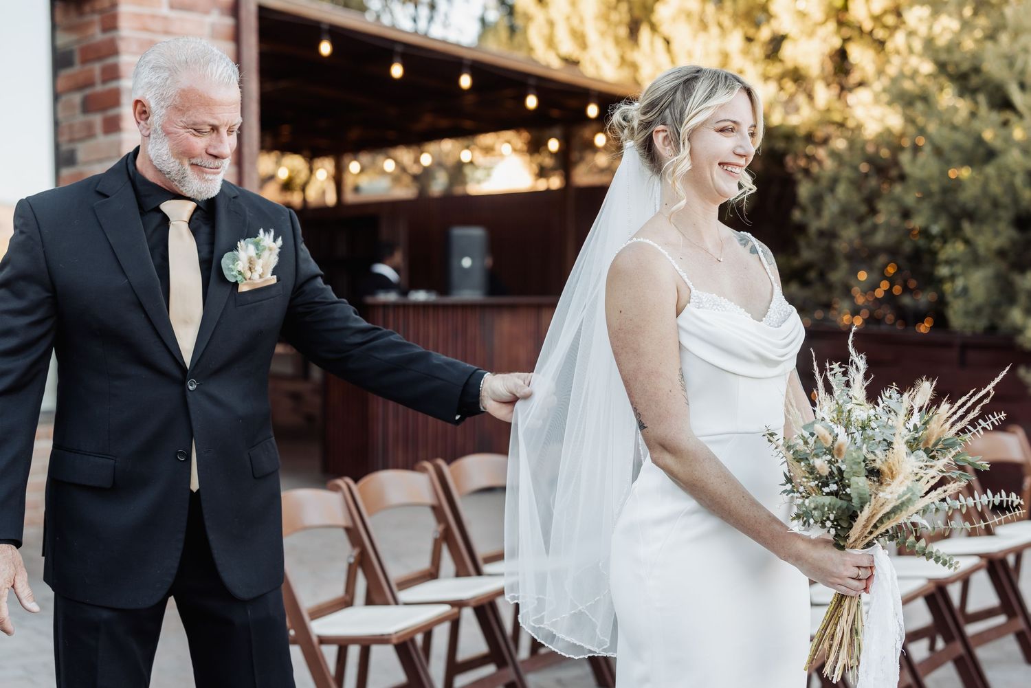 A bride in a white dress holding a bouquet stands outdoors with companion in black tuxedo at wedding ceremony setup.