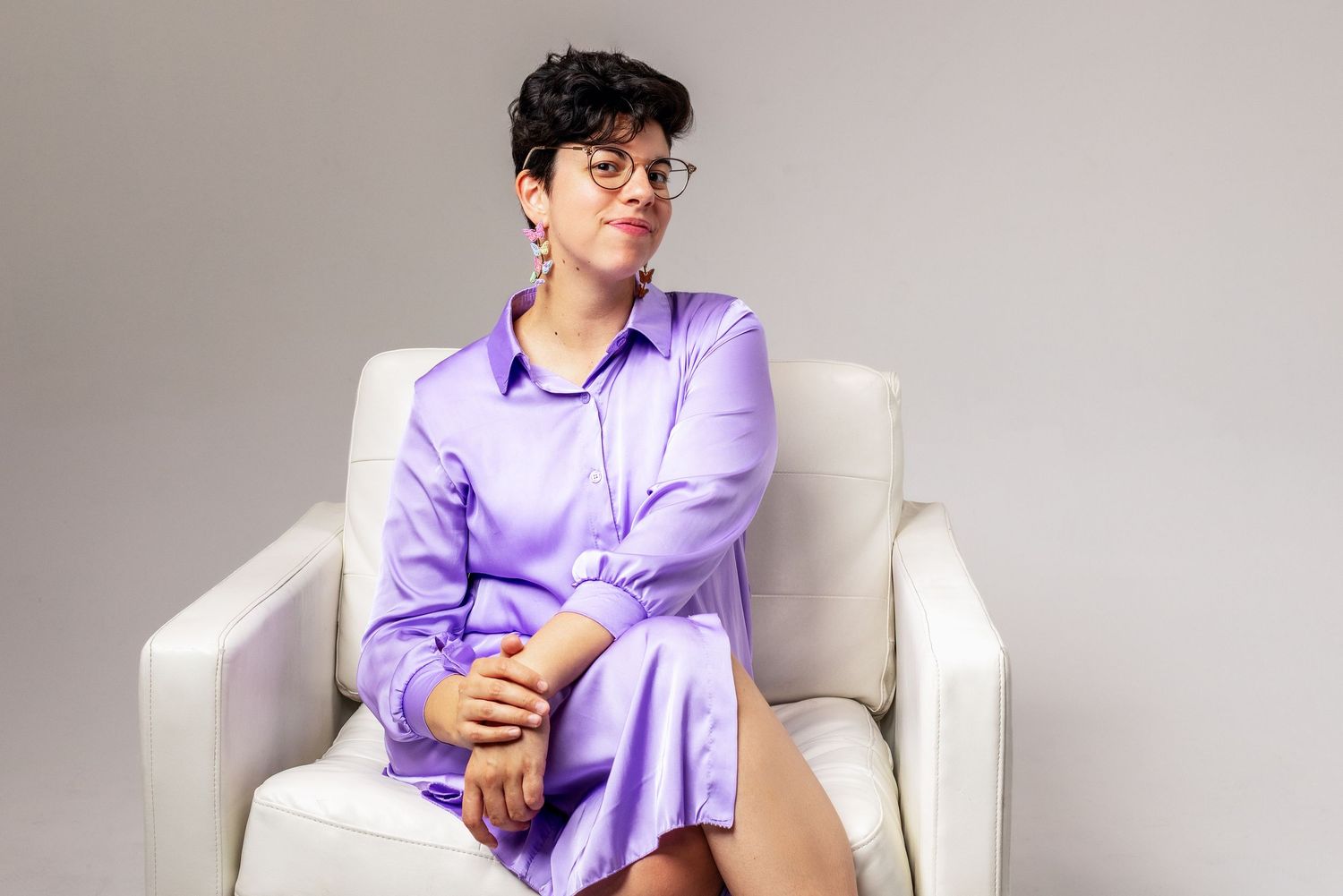 A person in a lilac button-down outfit sits in a white armchair against a neutral backdrop.