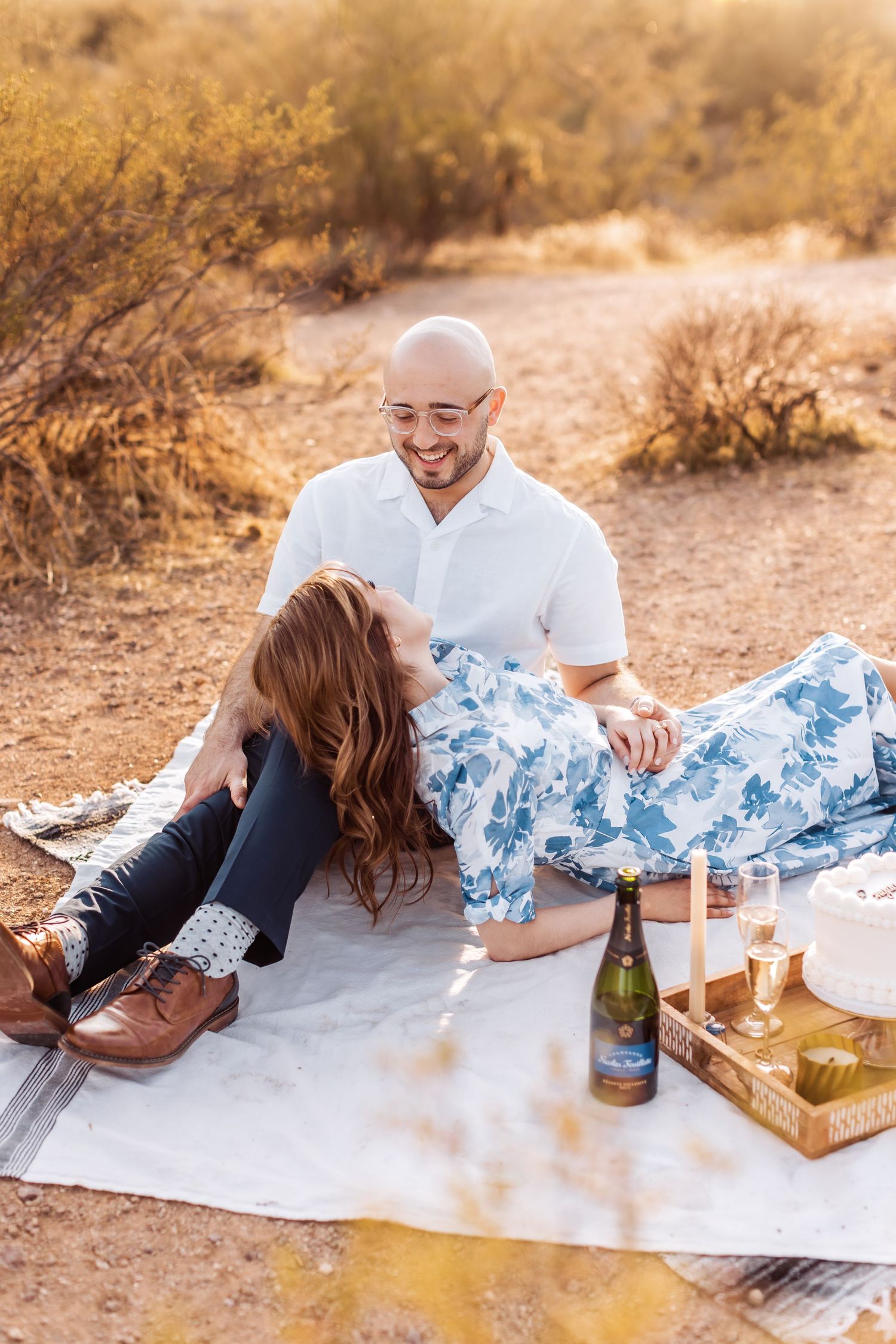 A couple enjoys a romantic desert picnic with wine and candles at sunset against rugged terrain.