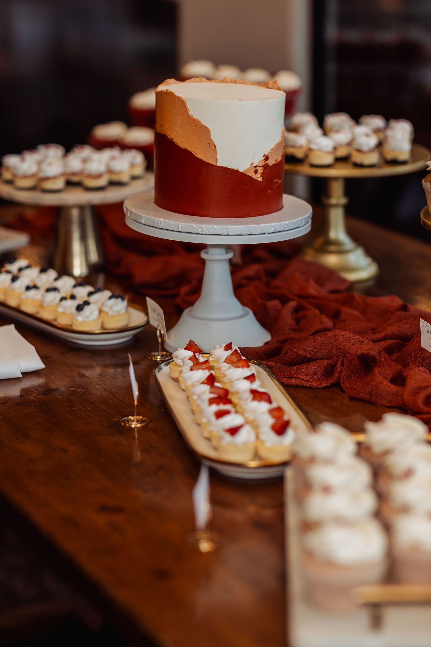 A rustic dessert table display featuring a white cake on a pedestal and various mini desserts arranged on dark wood.