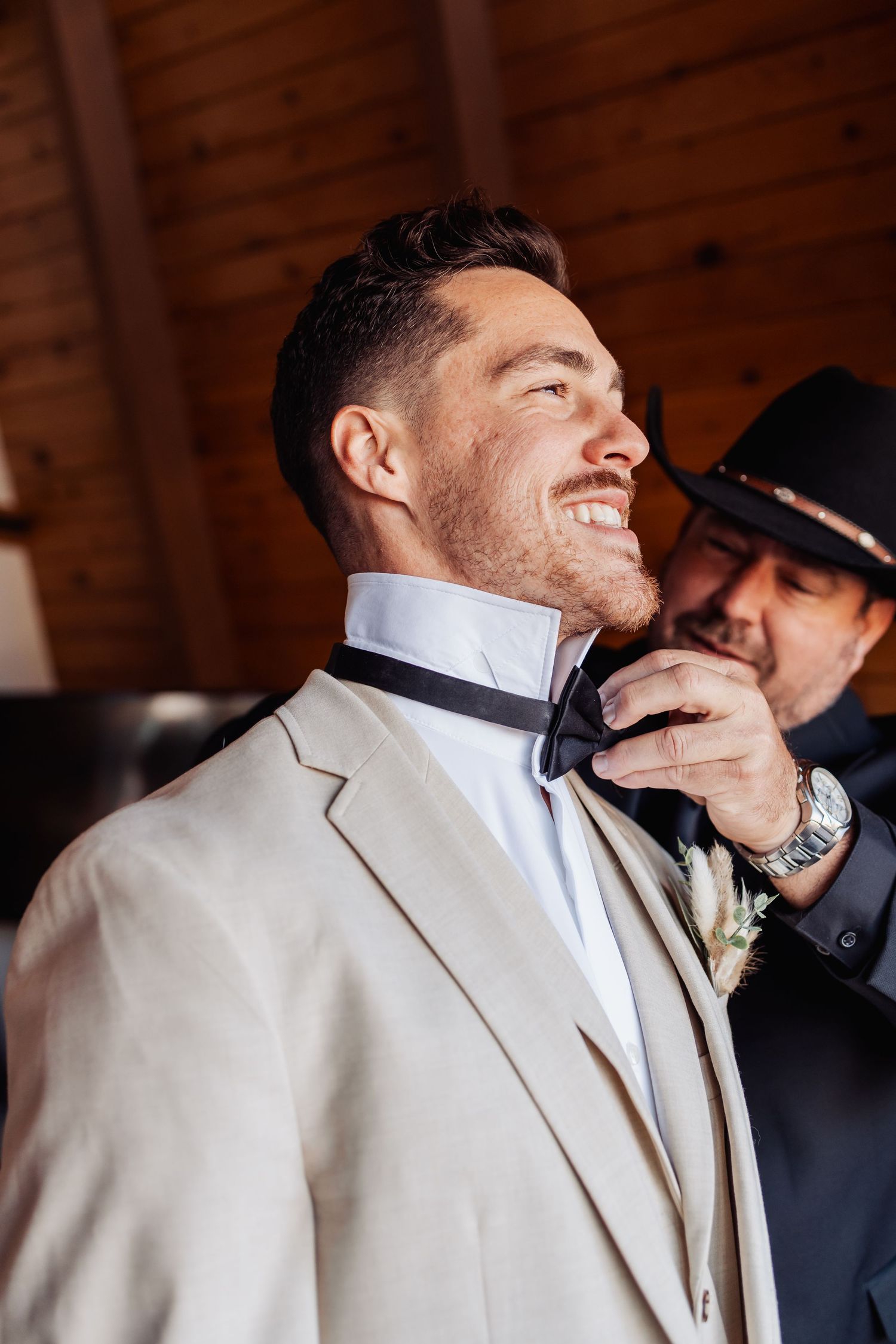Groom getting his bow tie adjusted during wedding preparations inside a rustic wooden interior.