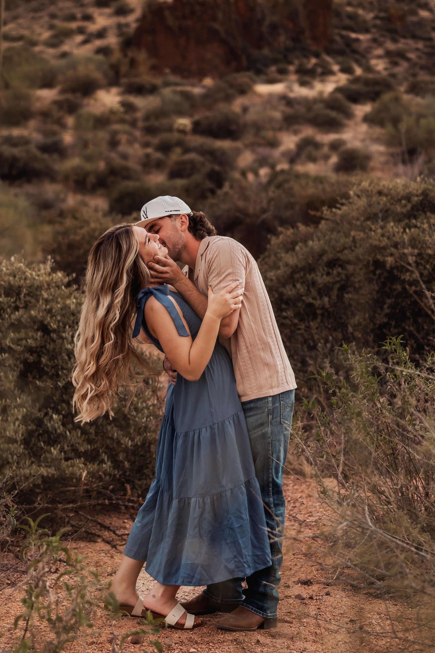 A couple shares a romantic kiss on a desert trail with rustic mountains in the background at sunset.