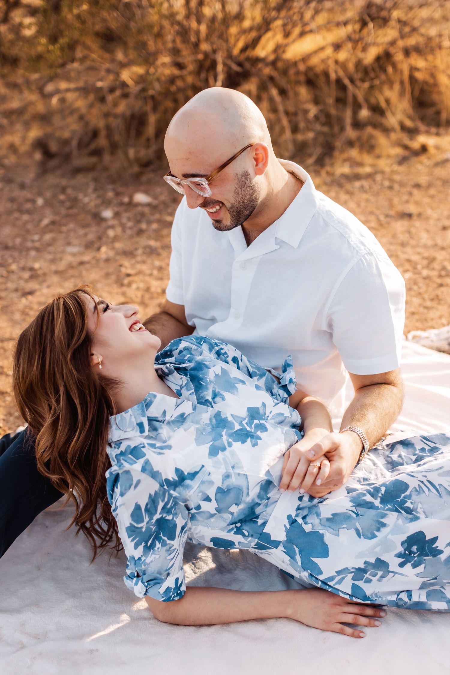 A couple shares a tender moment on a white blanket outdoors, with her wearing a blue floral dress and him in a white shirt.