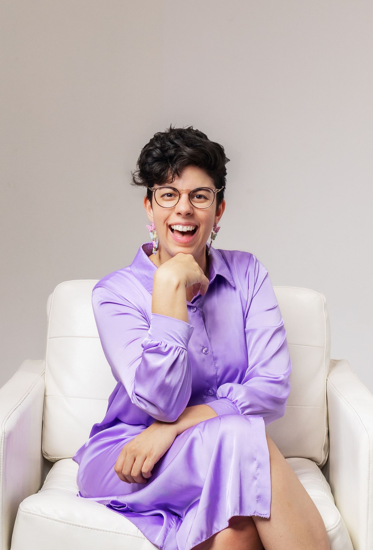 A person in round glasses and a purple dress sits on a white armchair with hand on chin while smiling enthusiastically.