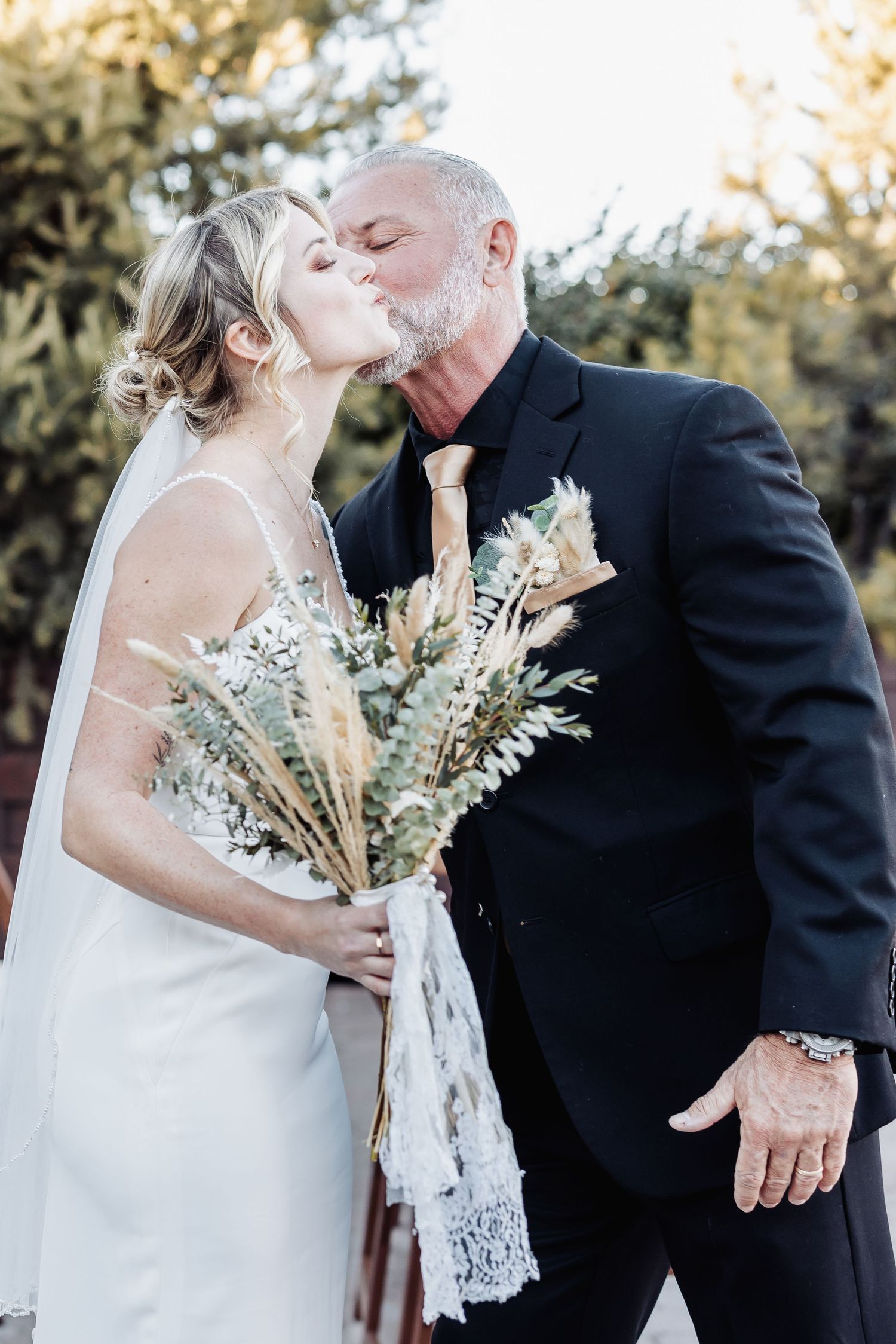 Newlyweds share a romantic kiss while holding a rustic bouquet of dried flowers and greenery.