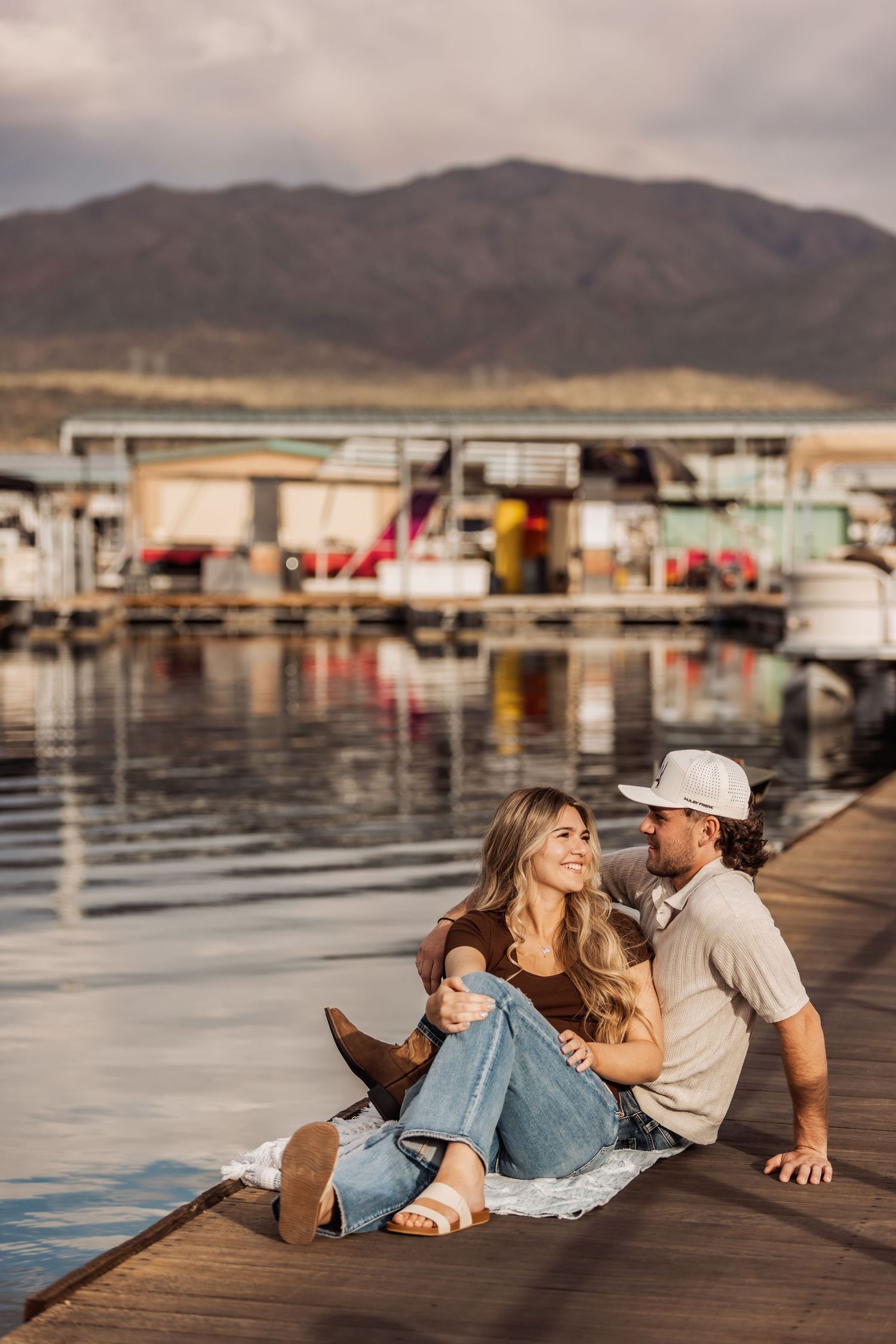 Romantic couple relaxes on wooden dock at marina with mountains and houseboats in background during sunset.
