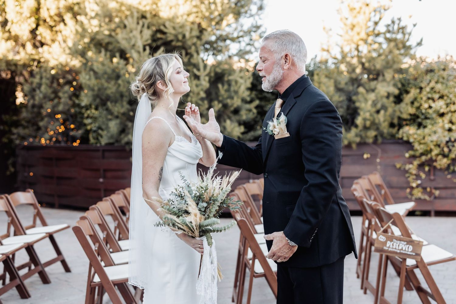 A tender moment between a couple at an outdoor wedding ceremony with wooden chairs and natural greenery in the background.