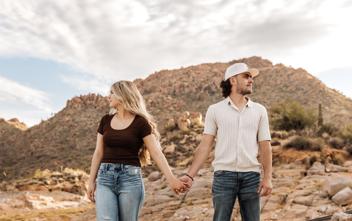 A couple holding hands while walking through a scenic desert landscape with rocky mountains behind them.